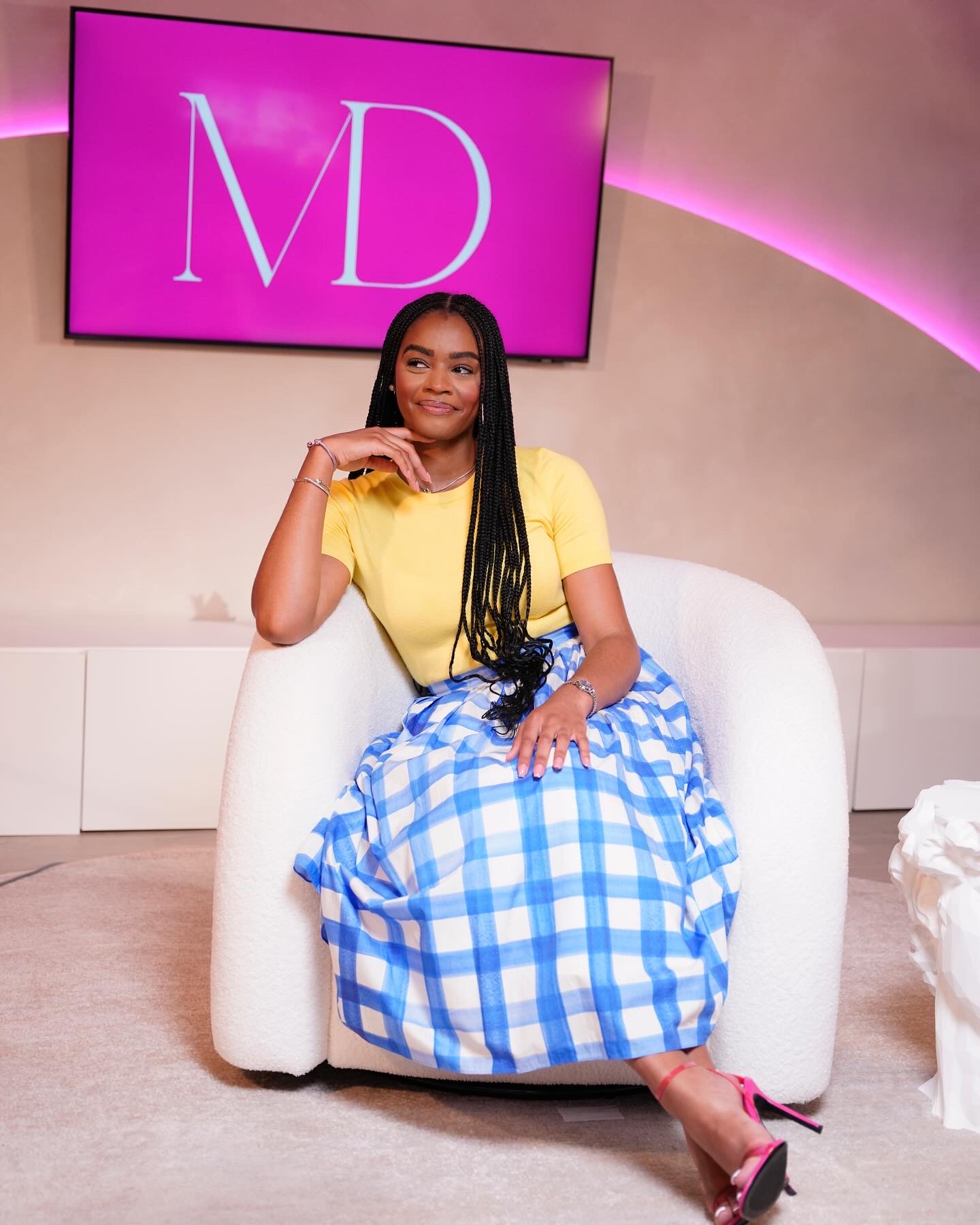 Woman sitting on white sofa in yellow top and blue checkered skirt, smiling, in studio with pink background and large screen displaying 'MD'.