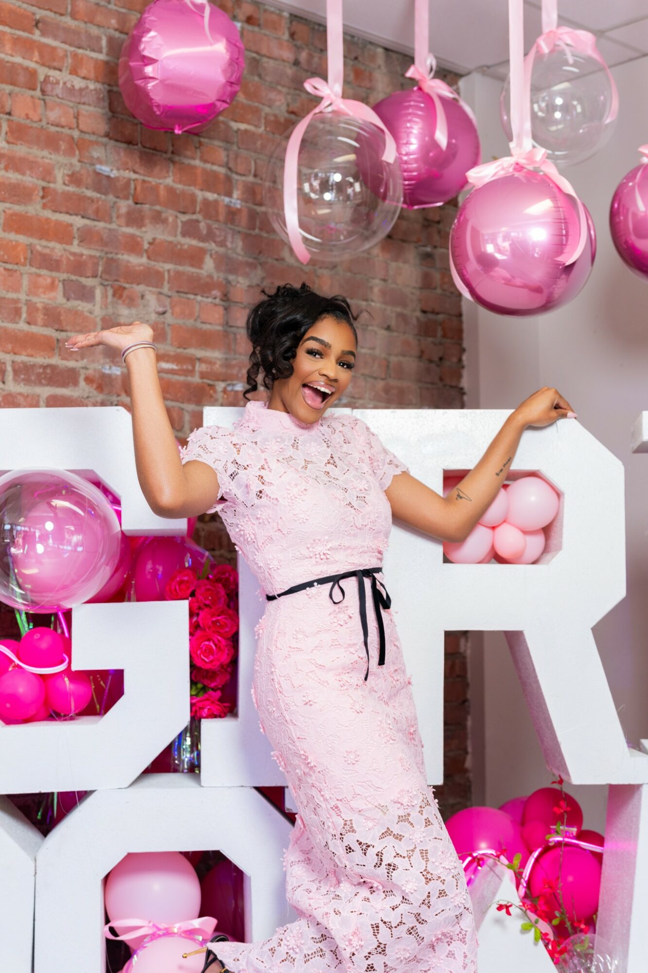 Woman in pink lace dress celebrating with balloons and decorations against brick wall.