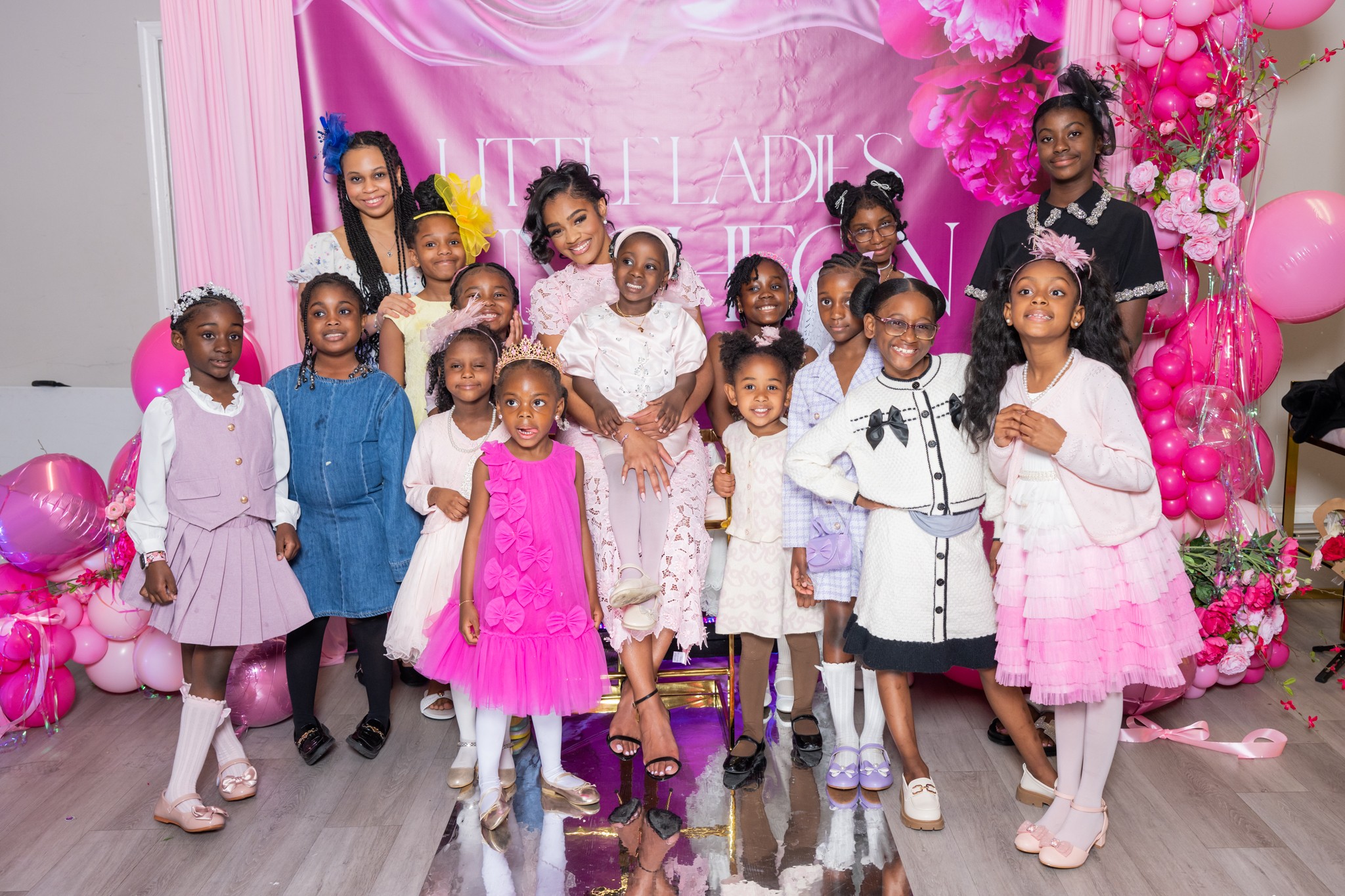 Group of children and adults celebrating with pink balloons and decorations, smiling in front of a pink backdrop.