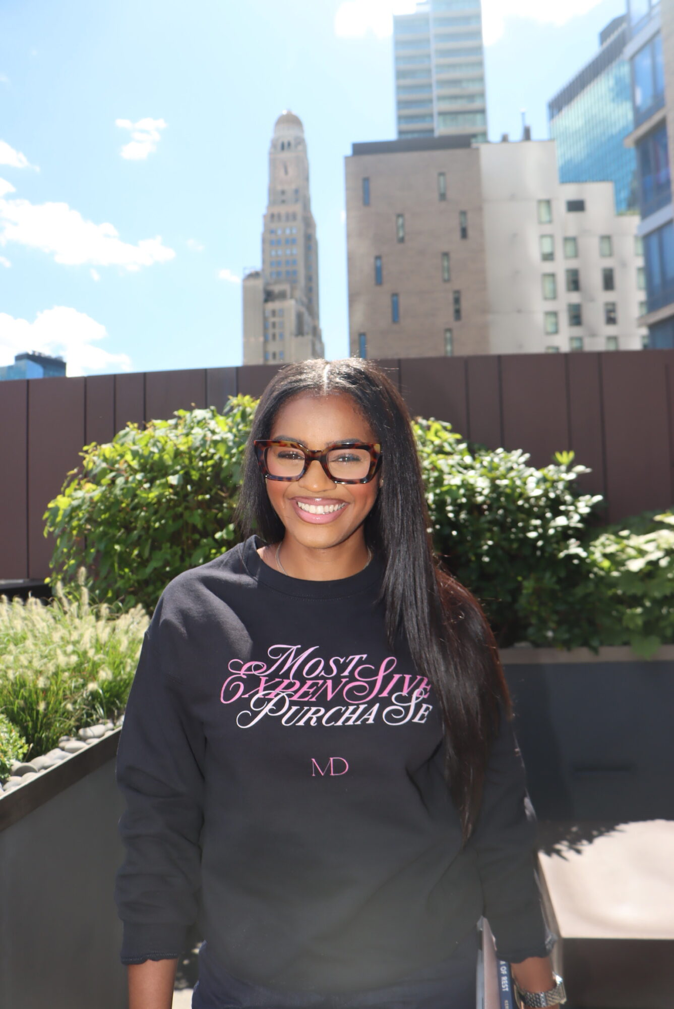 Smiling woman with glasses and long dark hair outdoors, city buildings in background, sunny day.