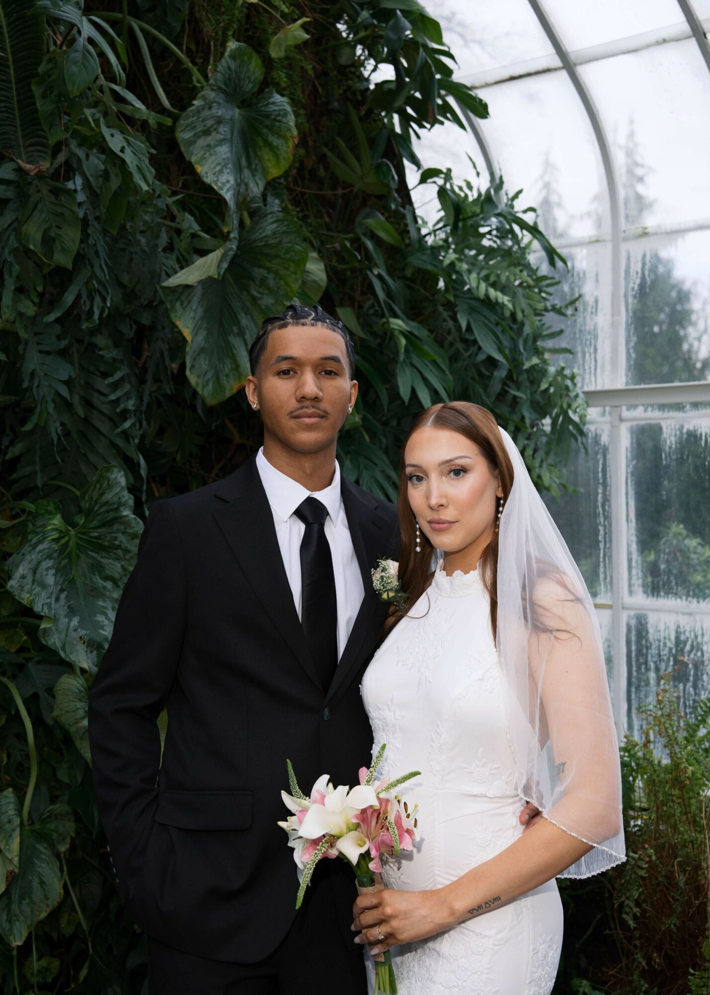 Bride and groom in wedding attire standing indoors with greenery and glass windows behind them.
