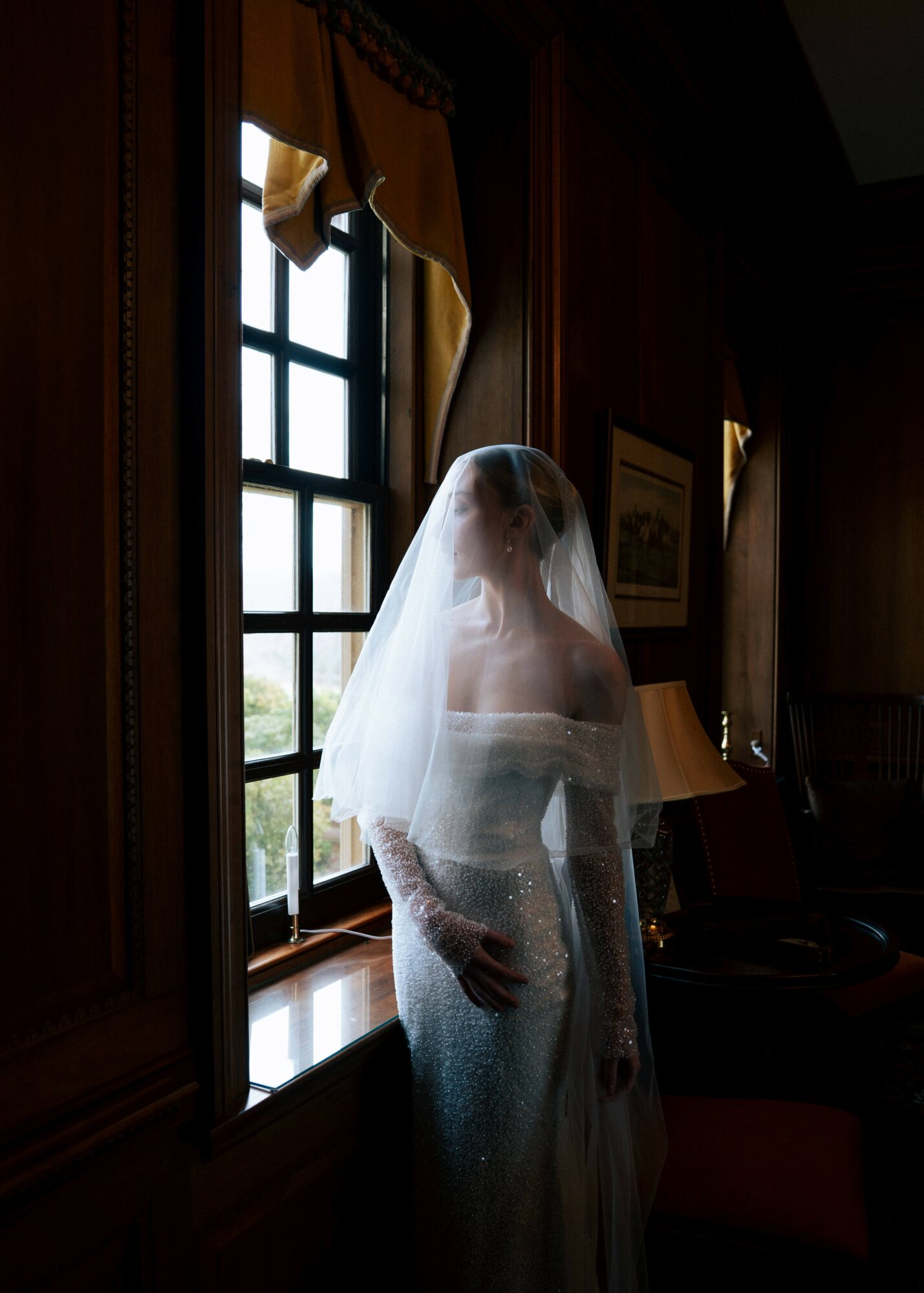 Person standing by window with curtain, wearing a veil, in a dimly lit room with wooden paneling and framed pictures.