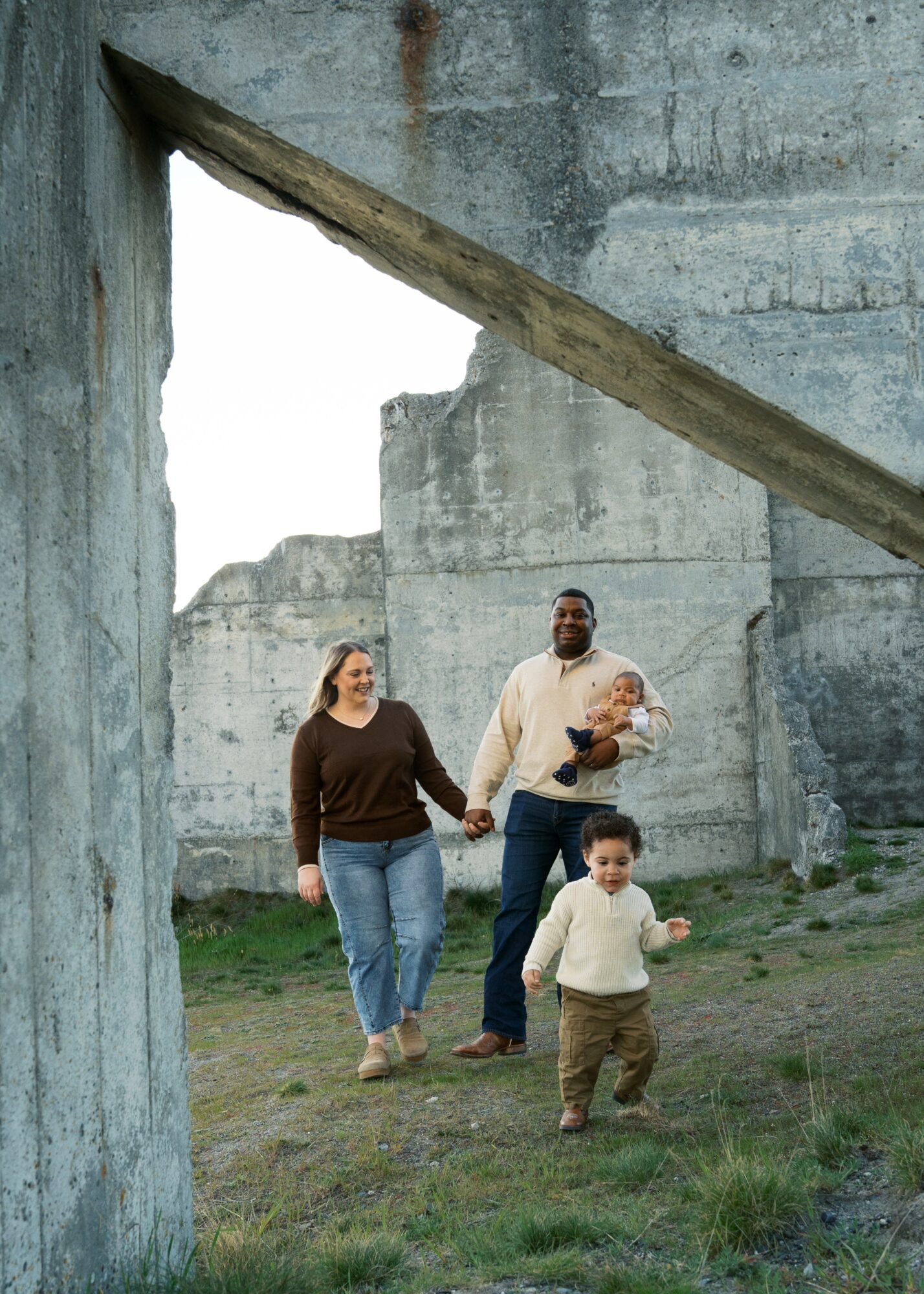 Four people, two adults and two children, walking outdoors near concrete structures, holding hands and smiling.
