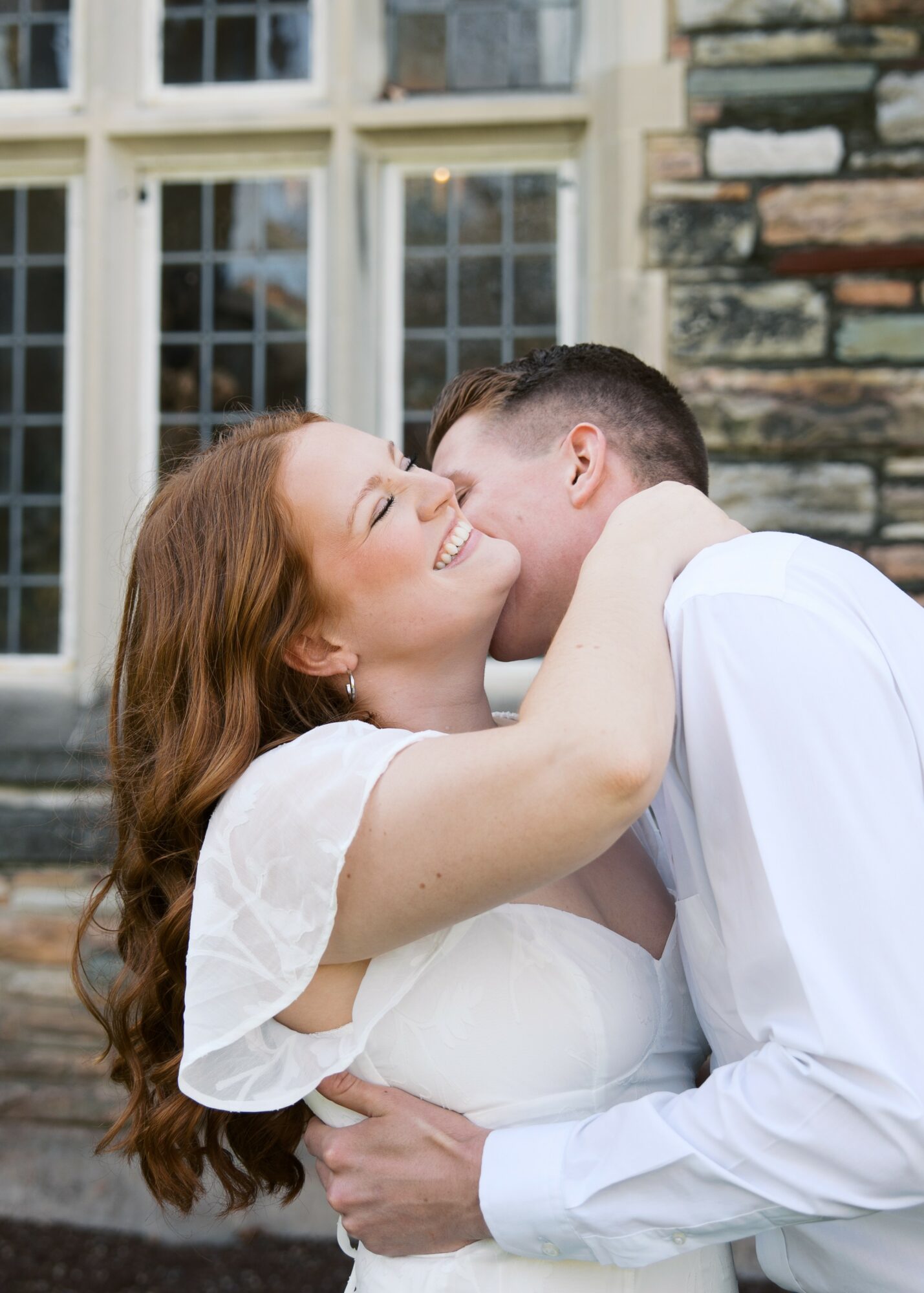 A couple embraces outdoors near a stone wall and large window, smiling and sharing a kiss.