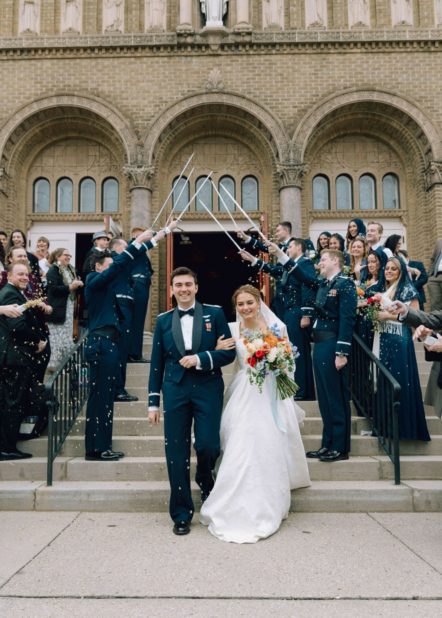 Bride and groom walking down stairs with wedding party and guests celebrating outside a church.