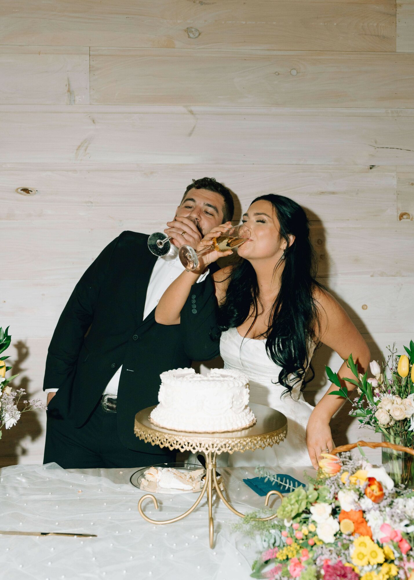Man and woman in formal attire sharing a slice of cake at a celebration, with a decorated table and floral arrangements.