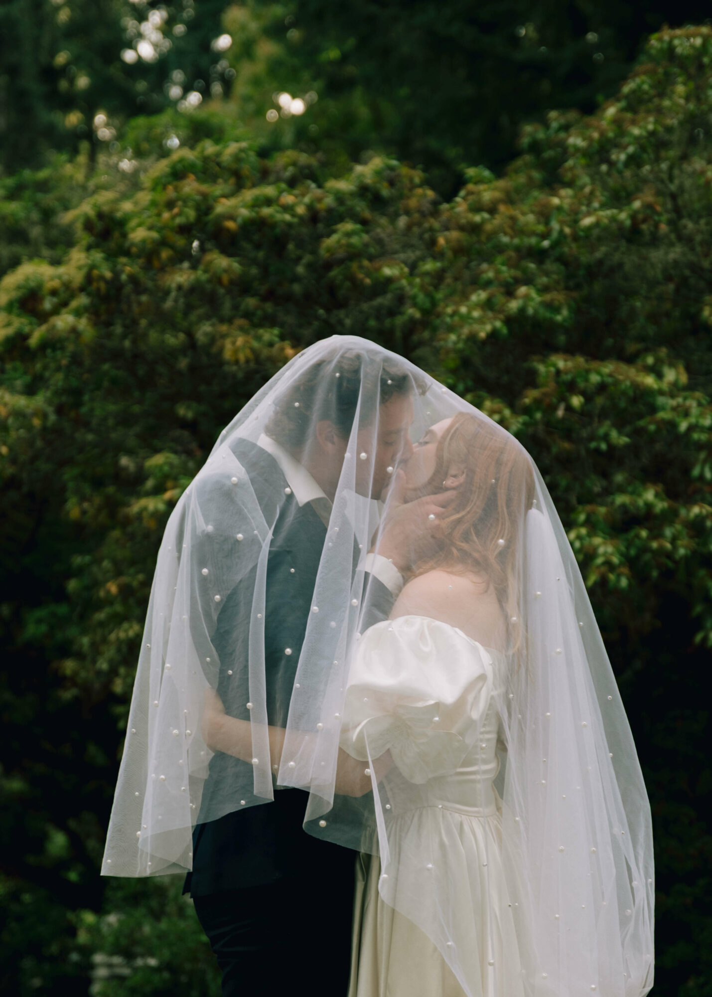Person wearing a wedding veil and white dress, standing outdoors with trees in background.