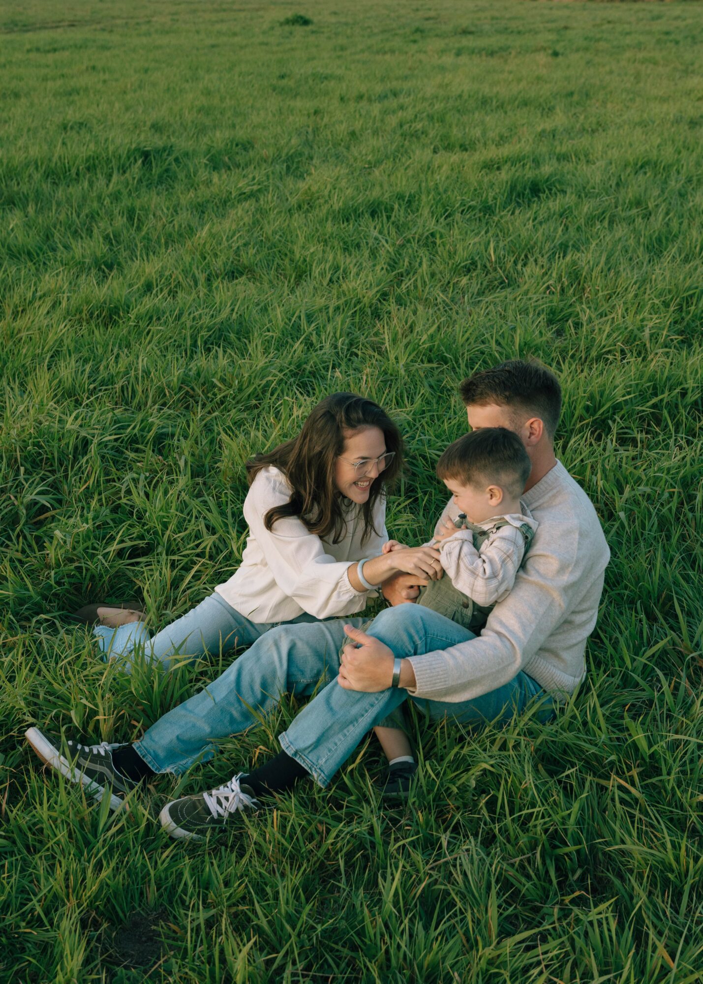 Family sitting on grass, playing and smiling outdoors in a green field.