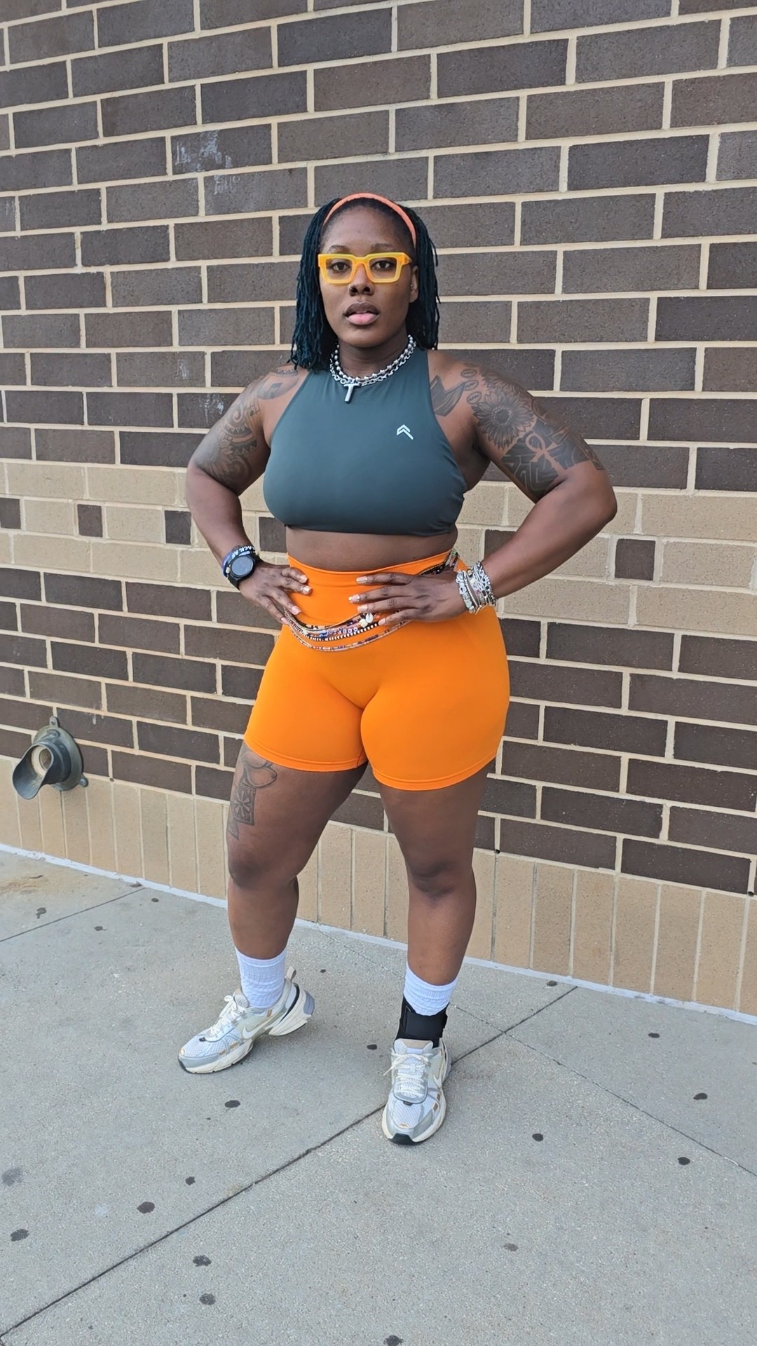 Person standing outdoors against a brick wall, wearing sunglasses, a sleeveless top, orange shorts, and sneakers.