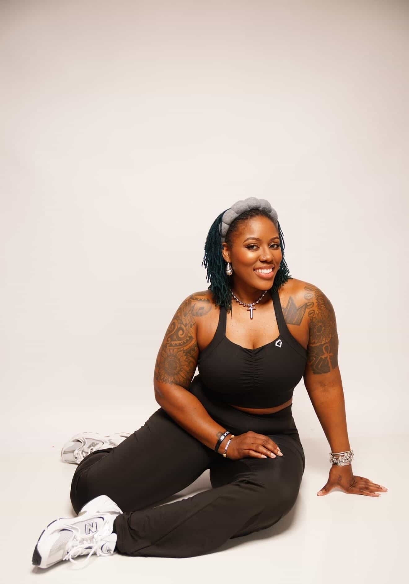 Woman with tattoos, wearing black tank top and pants, sitting on the floor against a plain background.