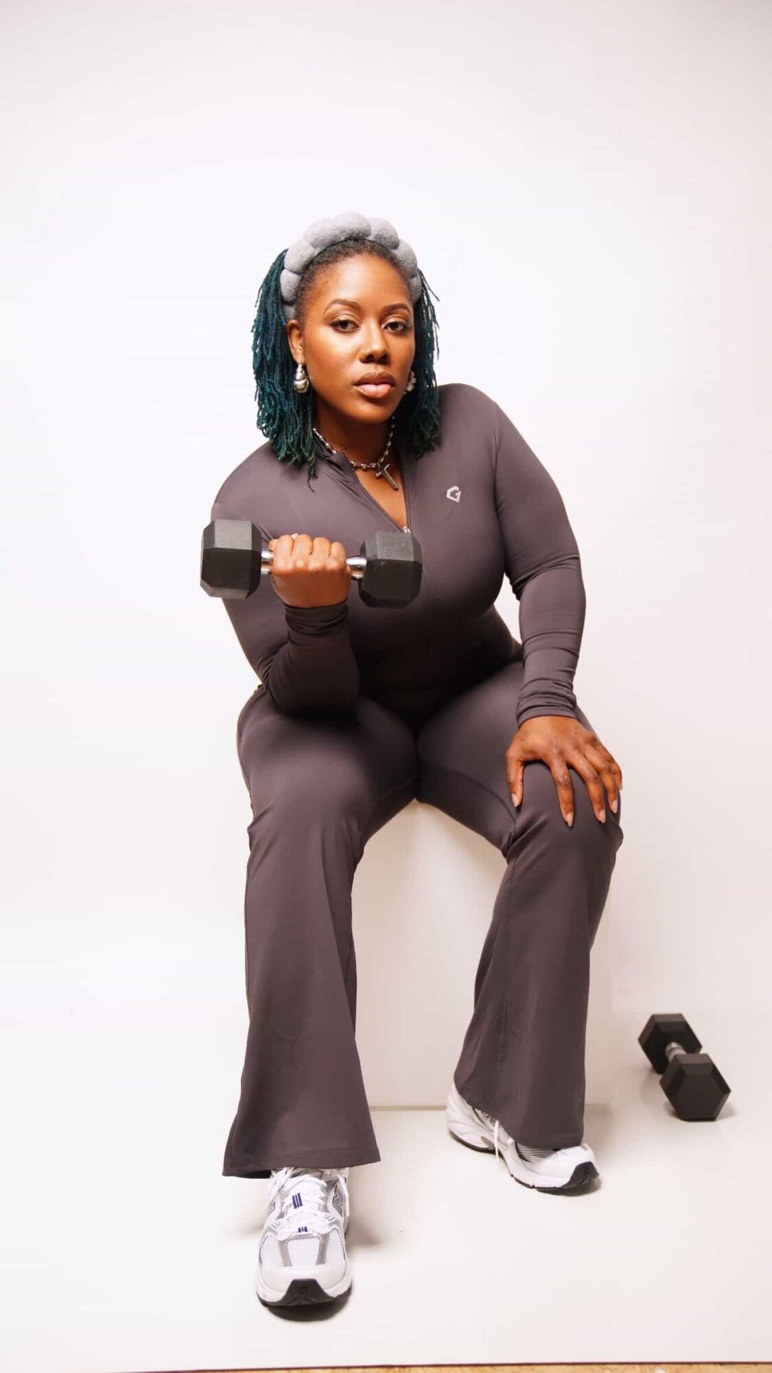 Woman sitting on a white surface holding a dumbbell, wearing dark clothing and sneakers, with a neutral background.