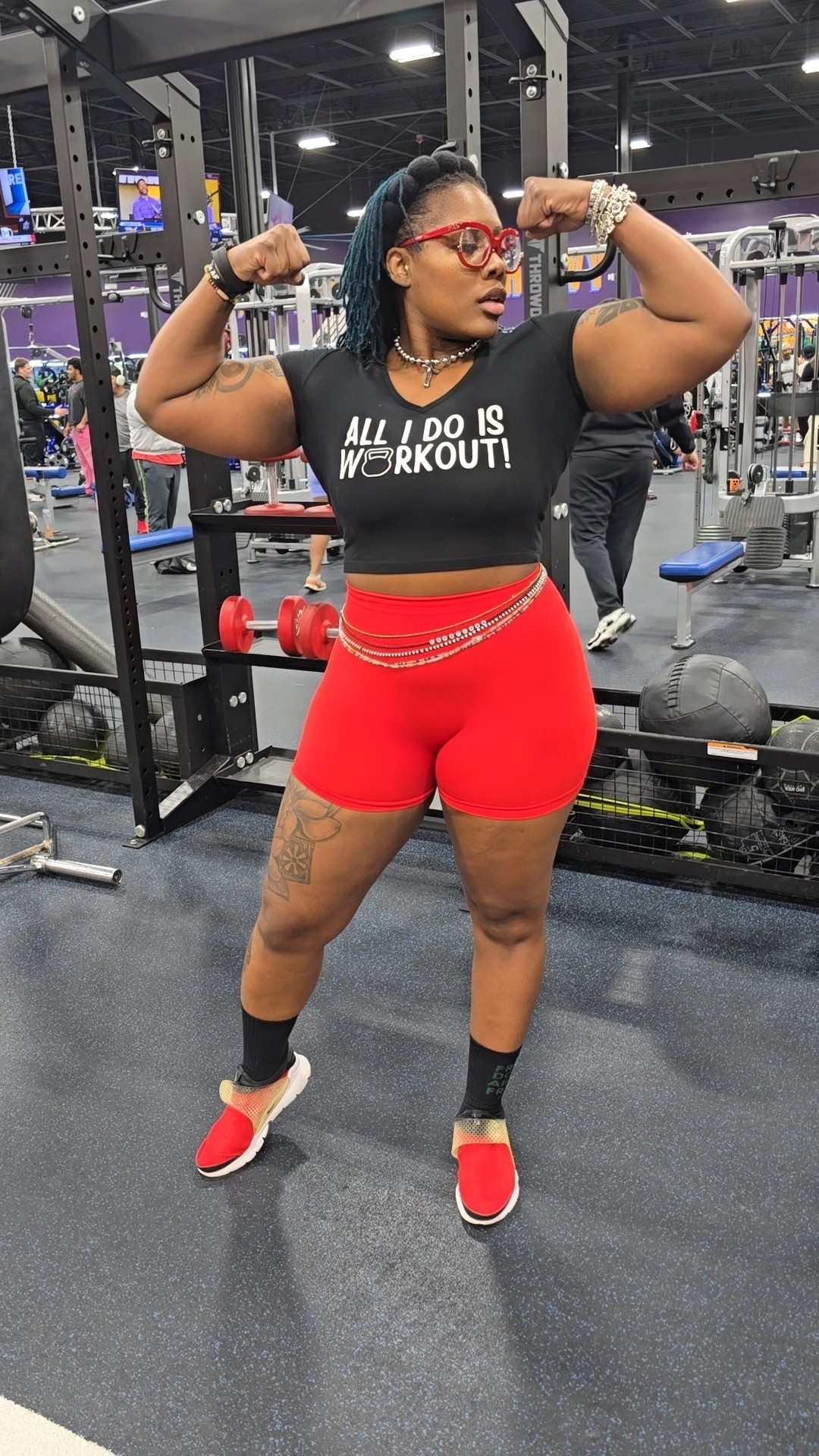 Woman in gym flexing arms, wearing black shirt, red shorts, glasses, and sneakers, with gym equipment in background.