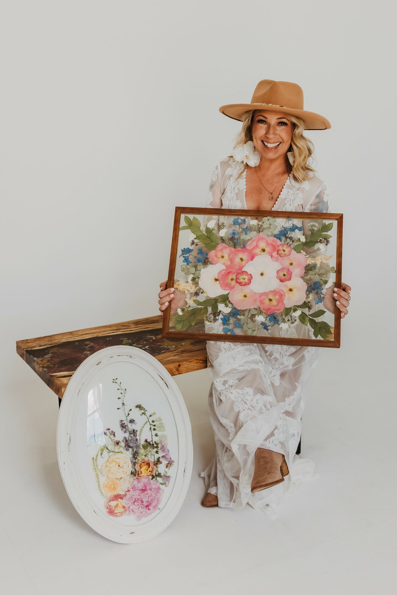 Woman with a hat holding a floral painting, sitting on the floor with a decorative plate nearby.