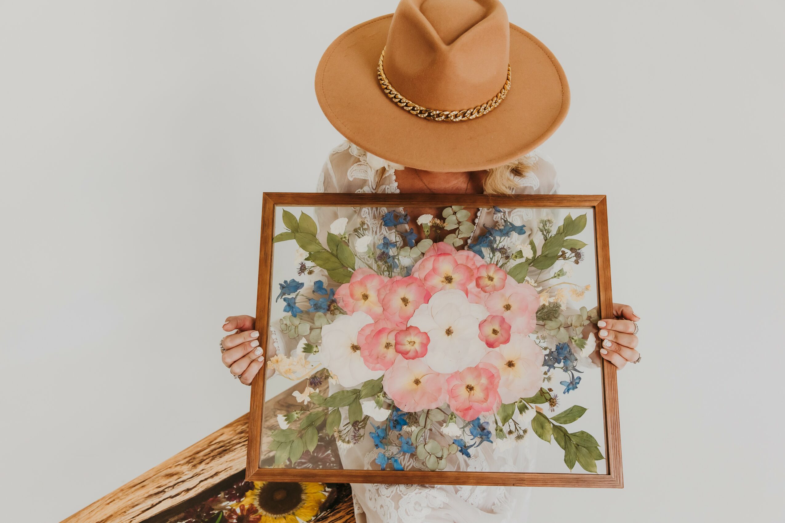 Person wearing a tan hat holding a framed floral painting with pink, blue, and green flowers.