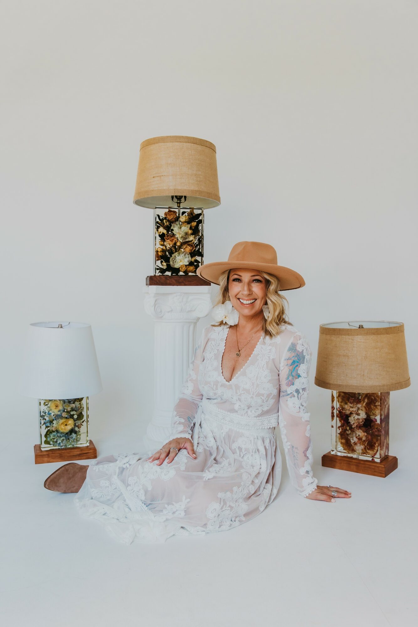 Woman in white dress and hat sitting on floor, surrounded by three lamps with floral bases, against plain background.