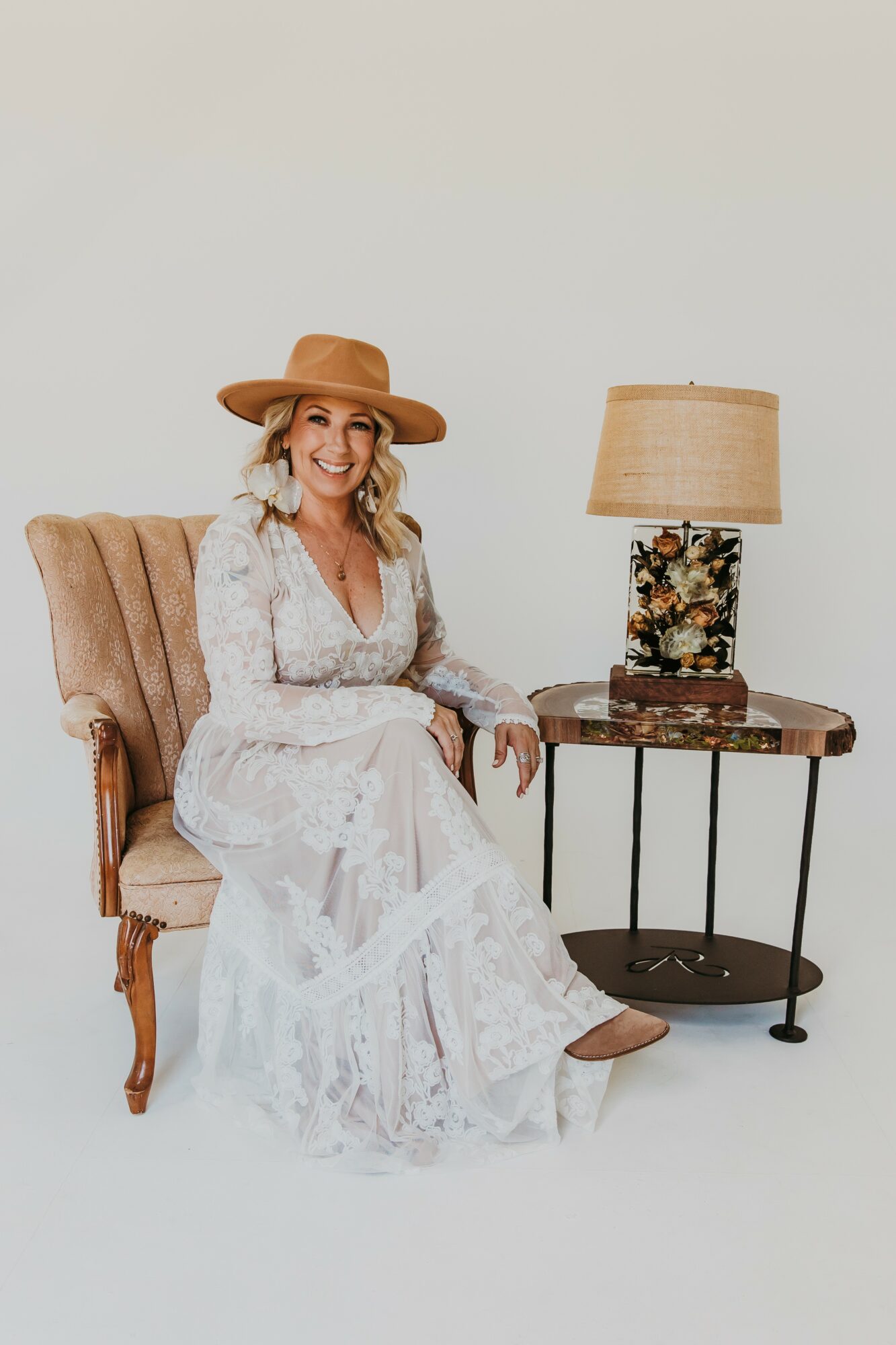 Woman in white dress and wide-brimmed hat sitting on a vintage armchair next to a side table with a lamp.