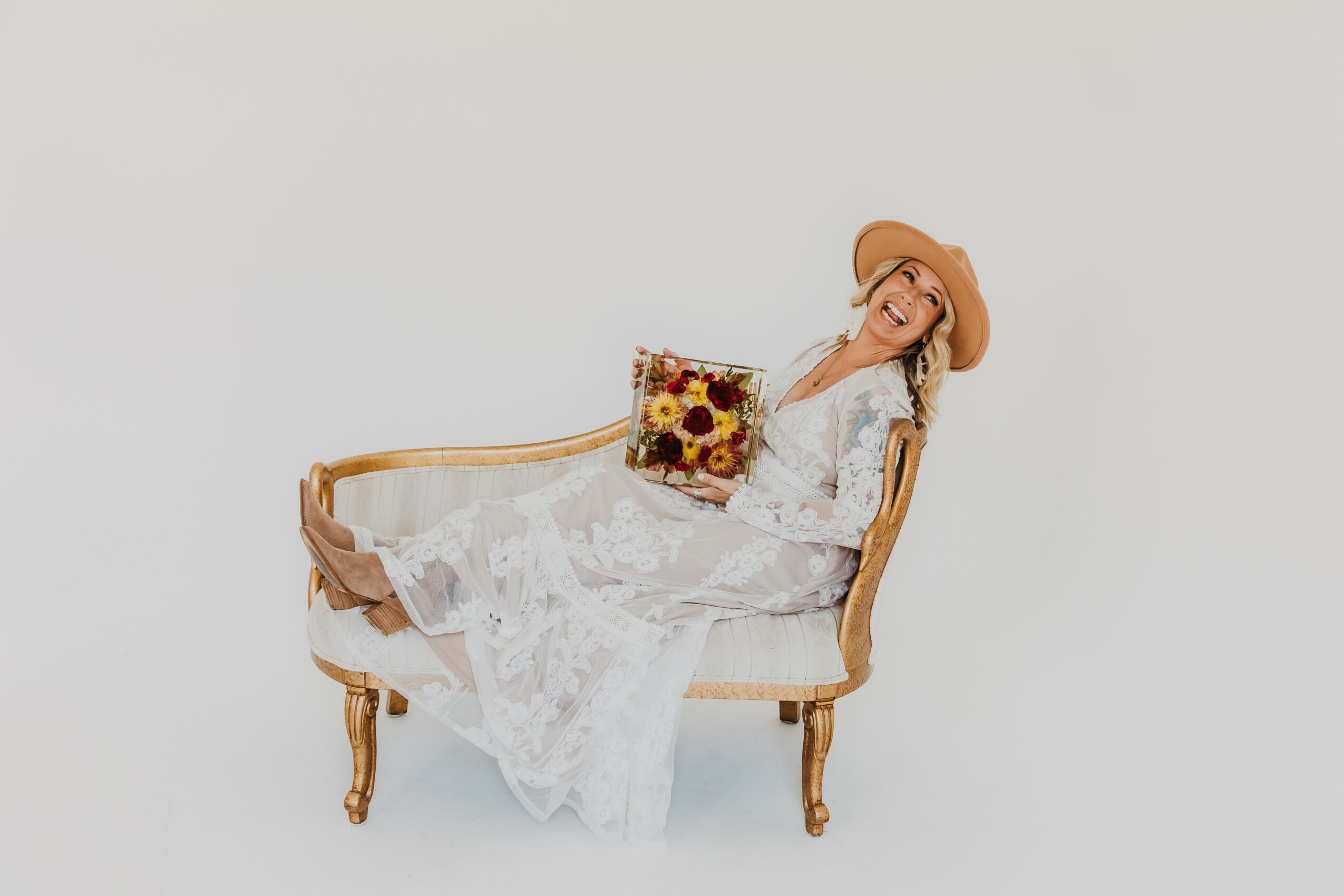Woman in a white lace dress and hat sitting on a vintage wooden bench, holding a sunflower-themed clutch.