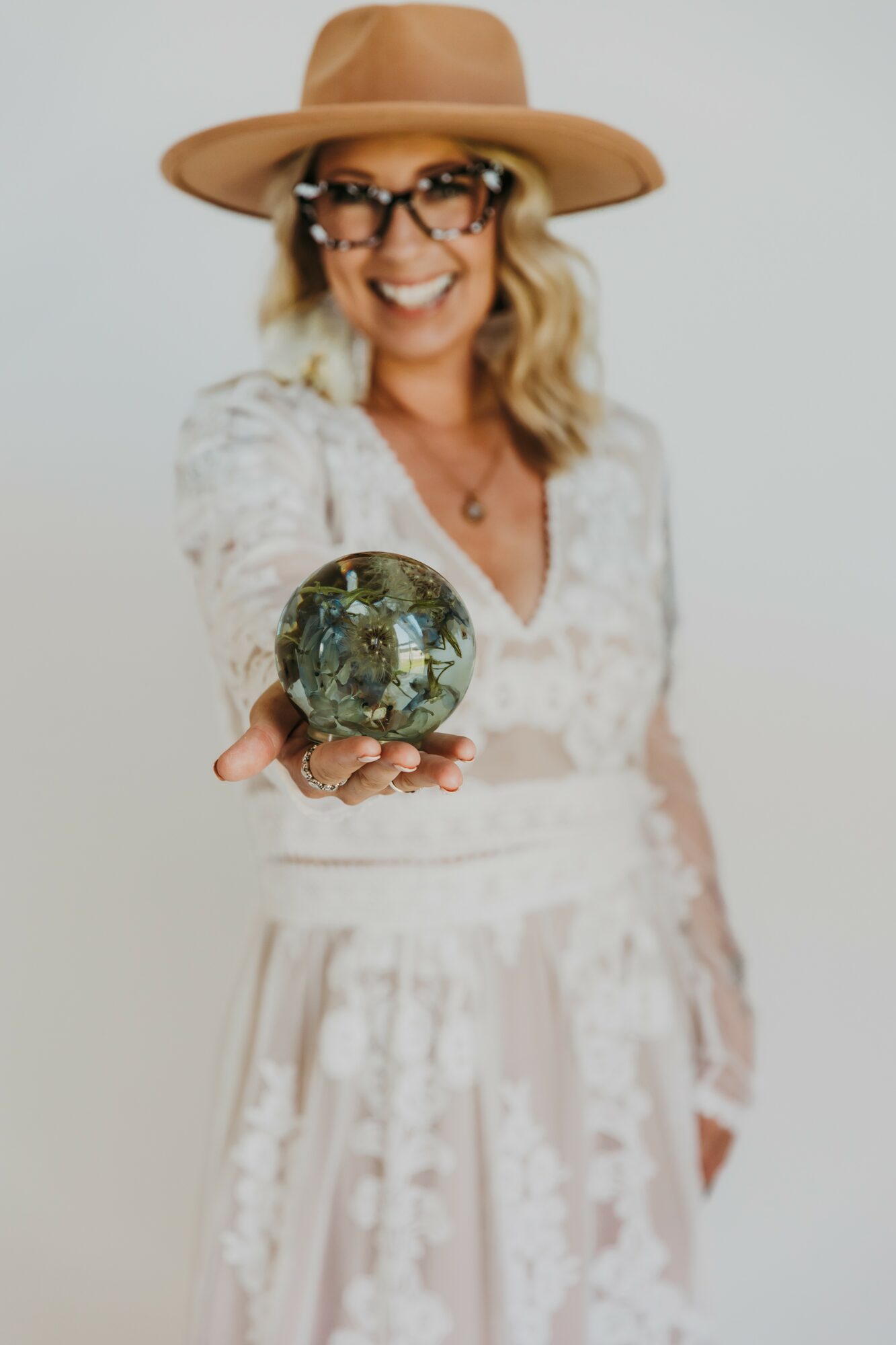 Woman wearing a hat and glasses holding a glass orb, smiling, in front of a plain background.