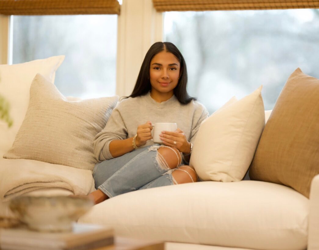 Young woman sitting on a sofa holding a mug, surrounded by cushions, in a bright room with large windows.