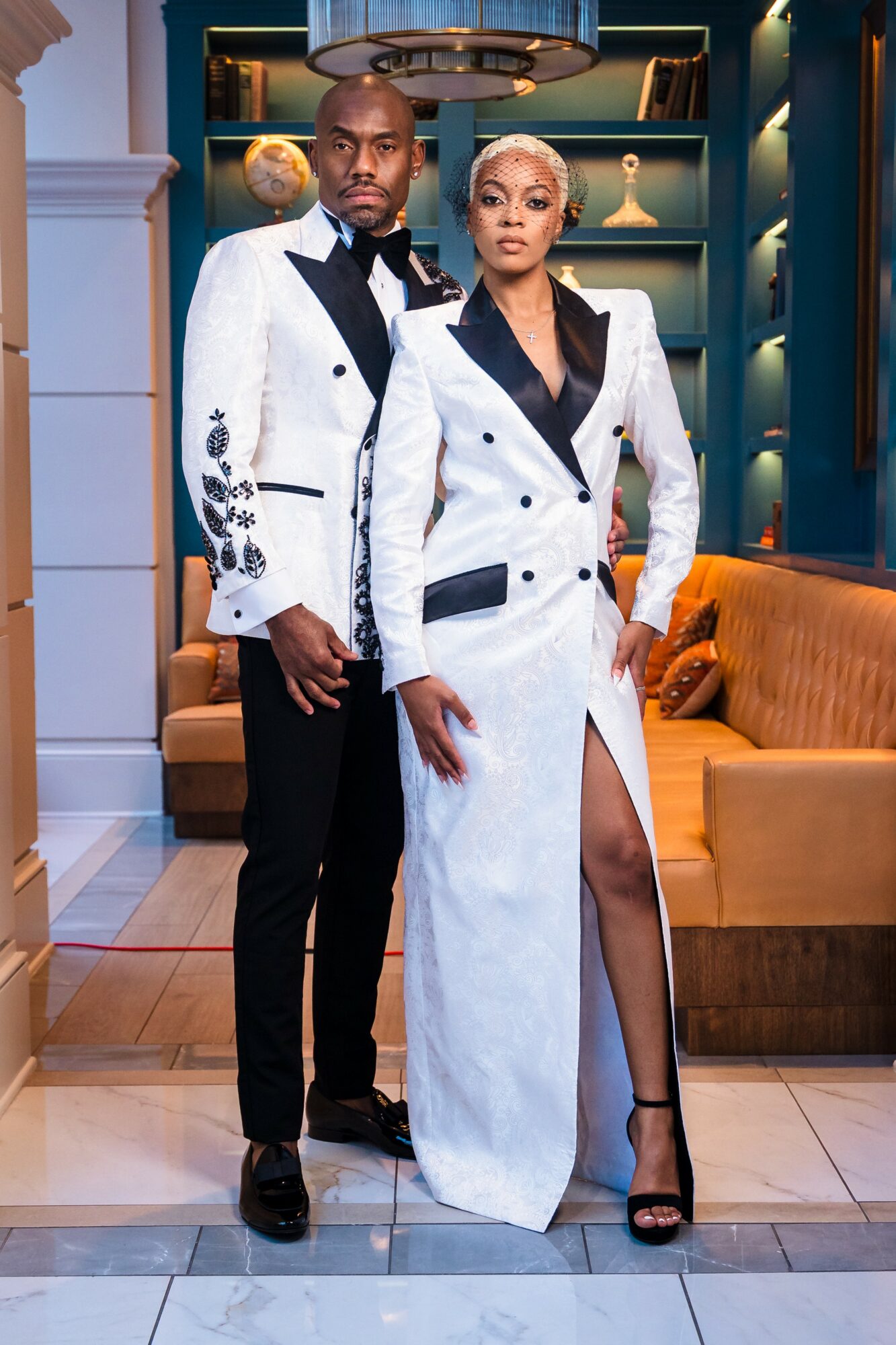 Man and woman in formal white and black outfits standing indoors with a sofa and shelves behind them.