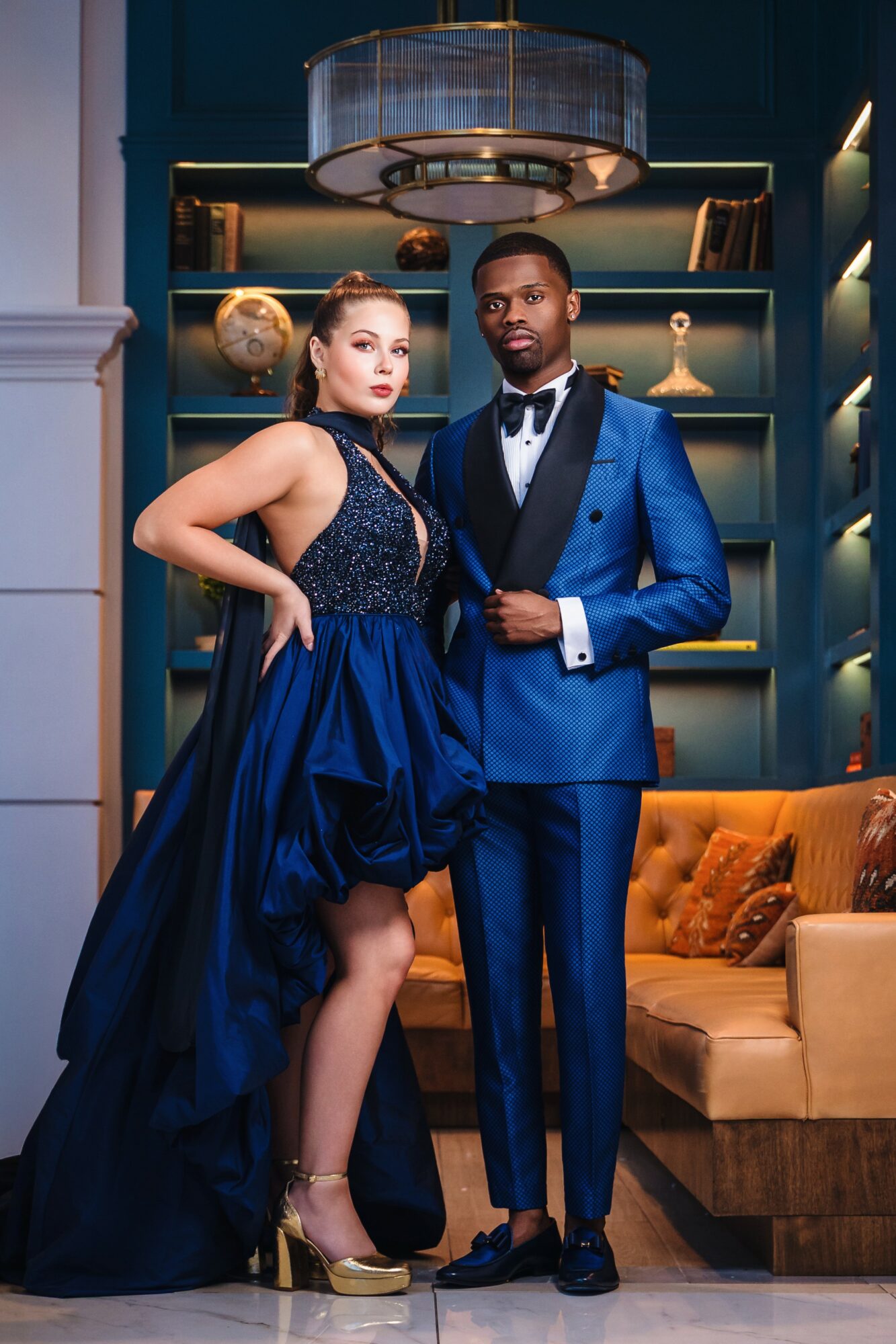 A woman and man in formal attire pose in a luxurious room with shelves and decorative objects.