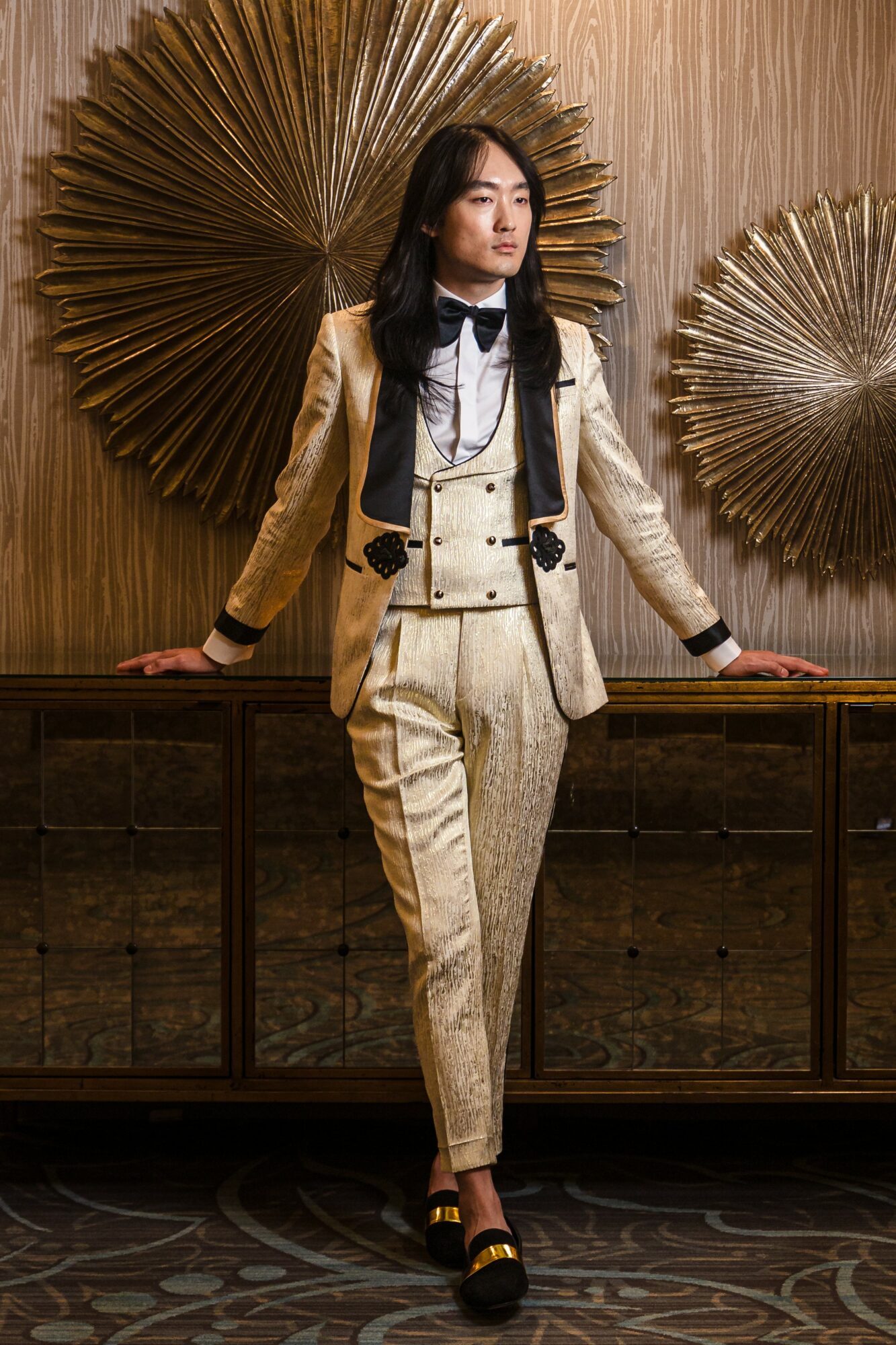 Woman in a beige suit with black accents, standing with hands on a wooden surface, decorative gold fan backdrop.