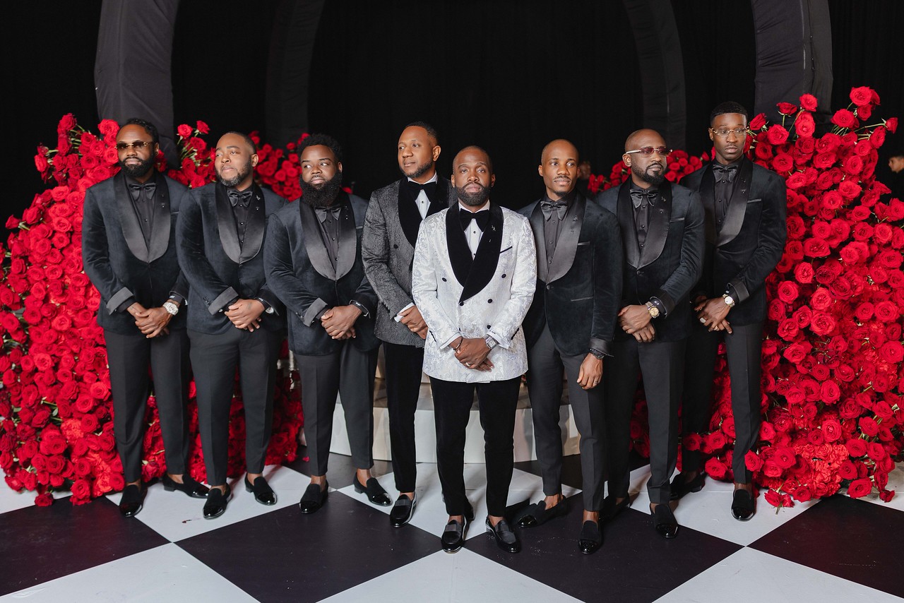 Group of eight men in formal suits standing on checkered floor with red floral backdrop.