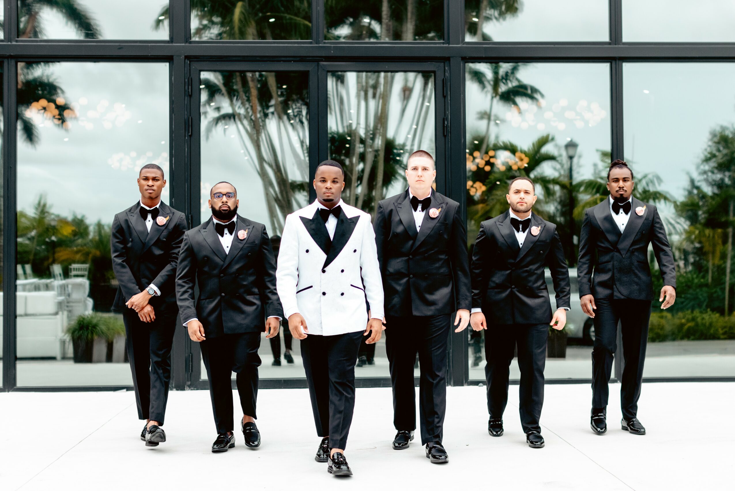 Six men in tuxedos walking outside in front of a glass building with trees reflected in the windows.
