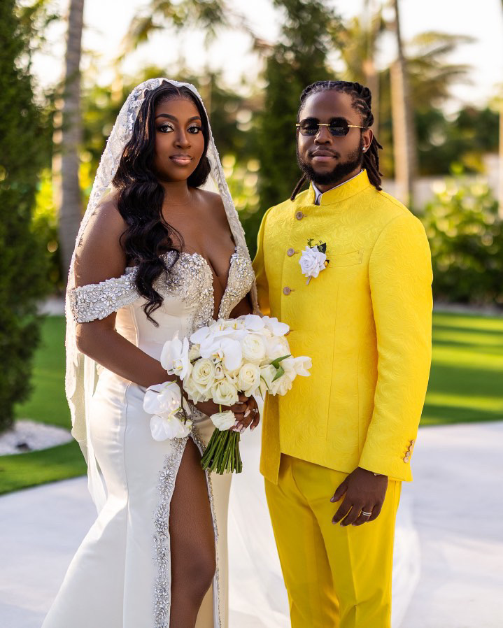 Bride in white dress holding a bouquet, groom in yellow suit with sunglasses outdoors, trees in background.