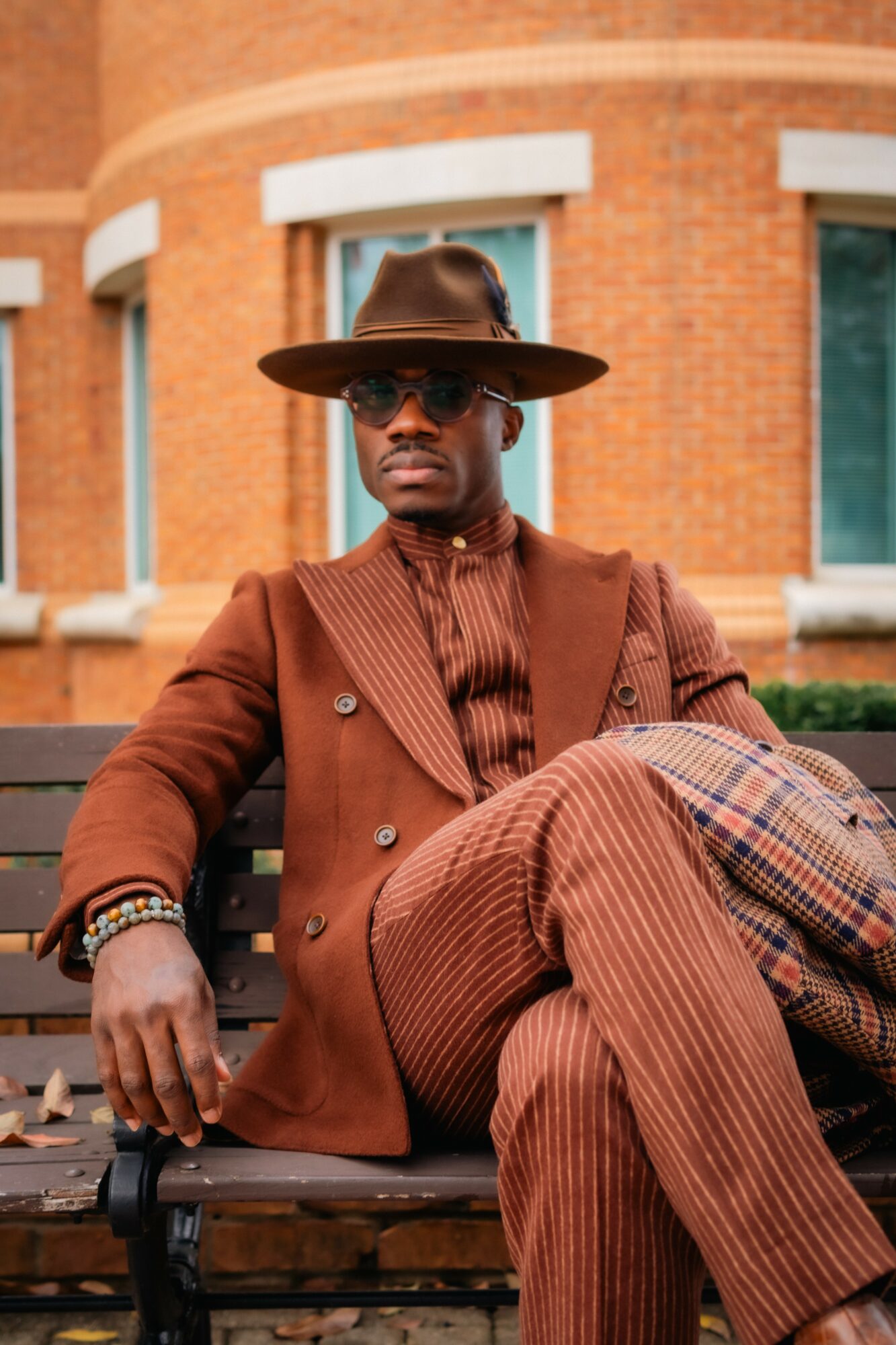 Man sitting on a bench wearing a wide-brimmed hat, sunglasses, and a brown suit with striped pants.