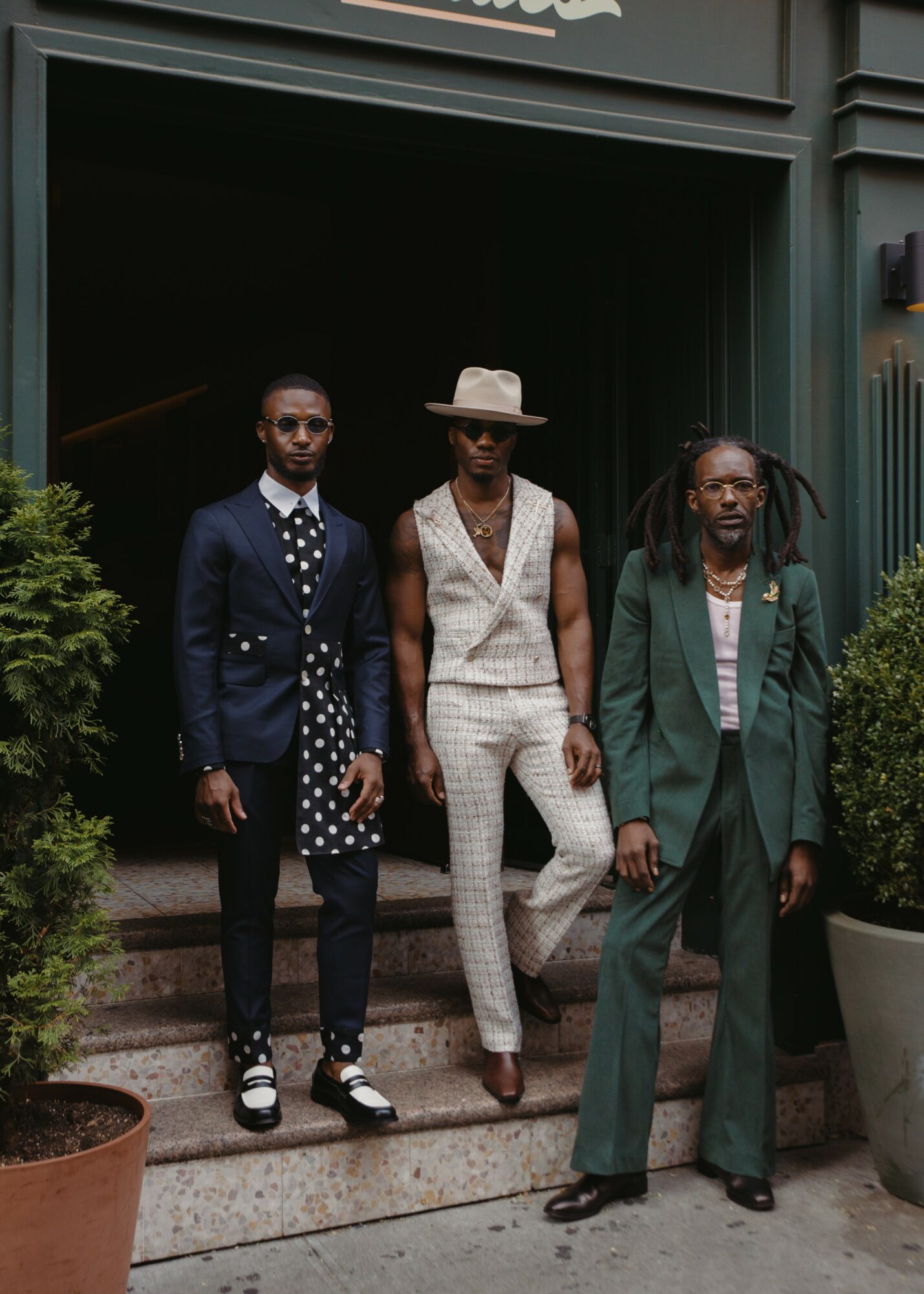 Three people standing on steps outside a building, dressed in formal and stylish clothing, with potted plants on sides.