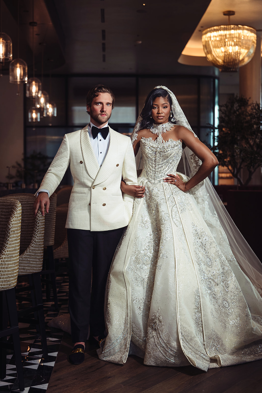 Bride and groom pose indoors, bride in a white wedding gown, groom in a tuxedo, elegant lighting and decor in background.