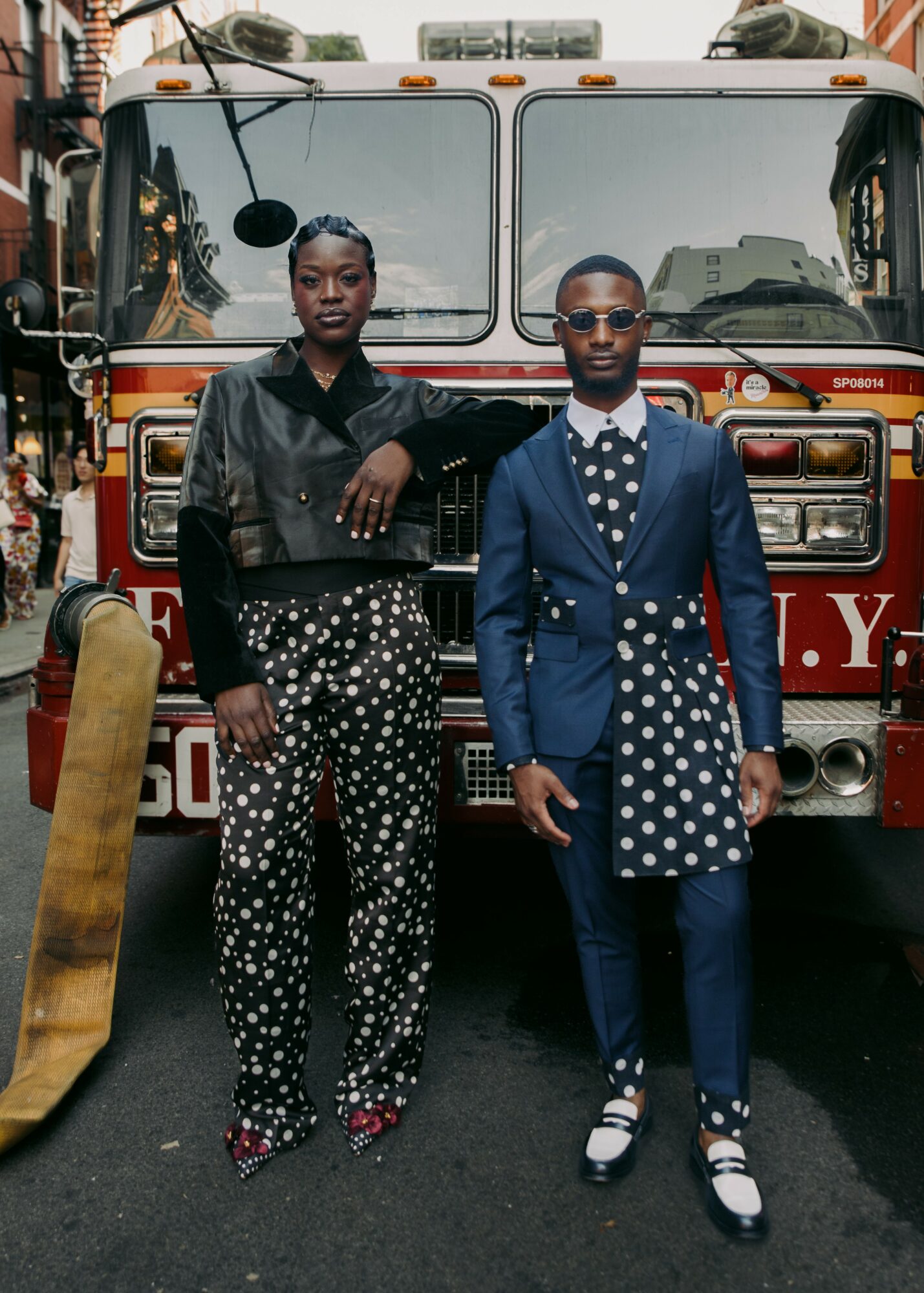 Two people stand in front of a fire truck on a city street, both dressed in stylish clothing.