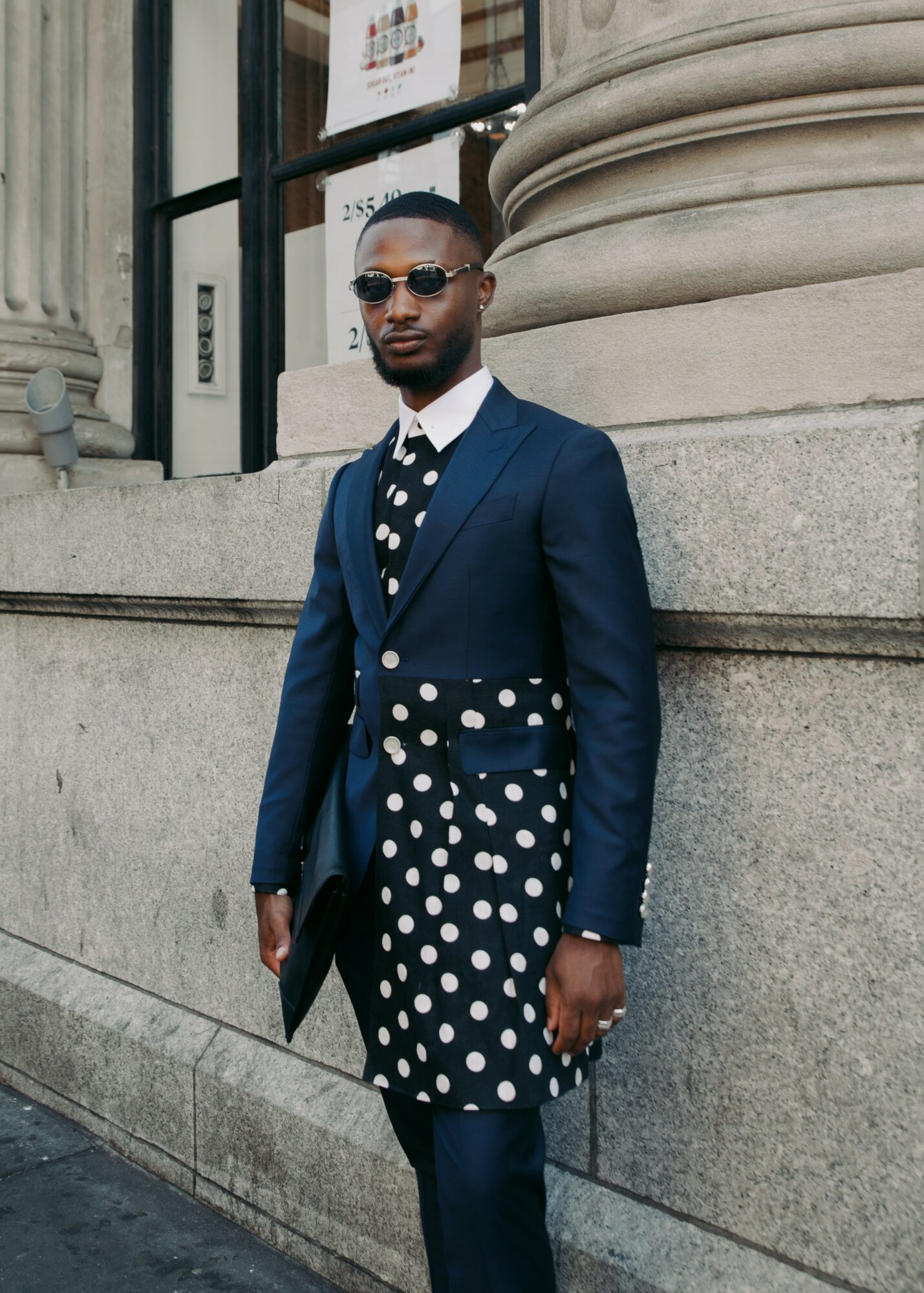 Man in a polka dot dress, navy blazer, and sunglasses standing outside a building with stone architecture.