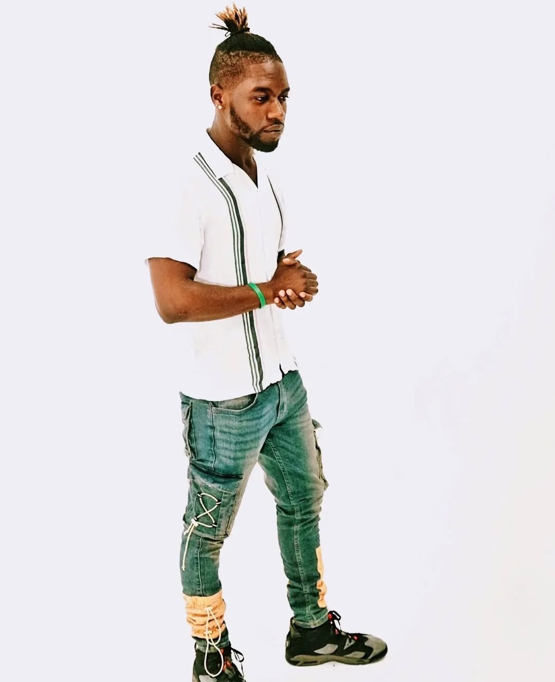 Man with a top knot hairstyle, wearing a white shirt and green jeans, standing with hands clasped, facing left.