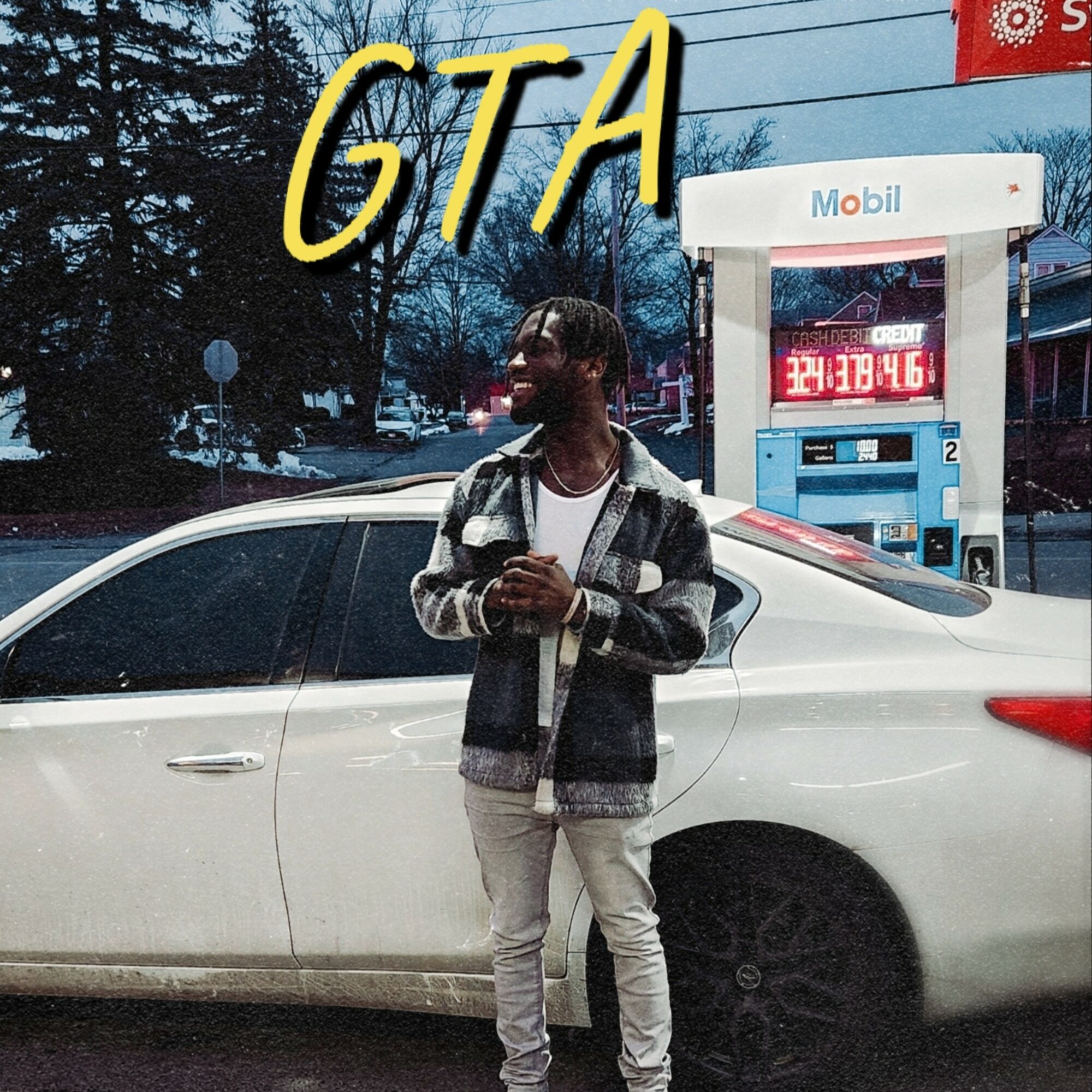 Person standing beside a silver car at a gas station, holding a phone, with trees and gas station in background.