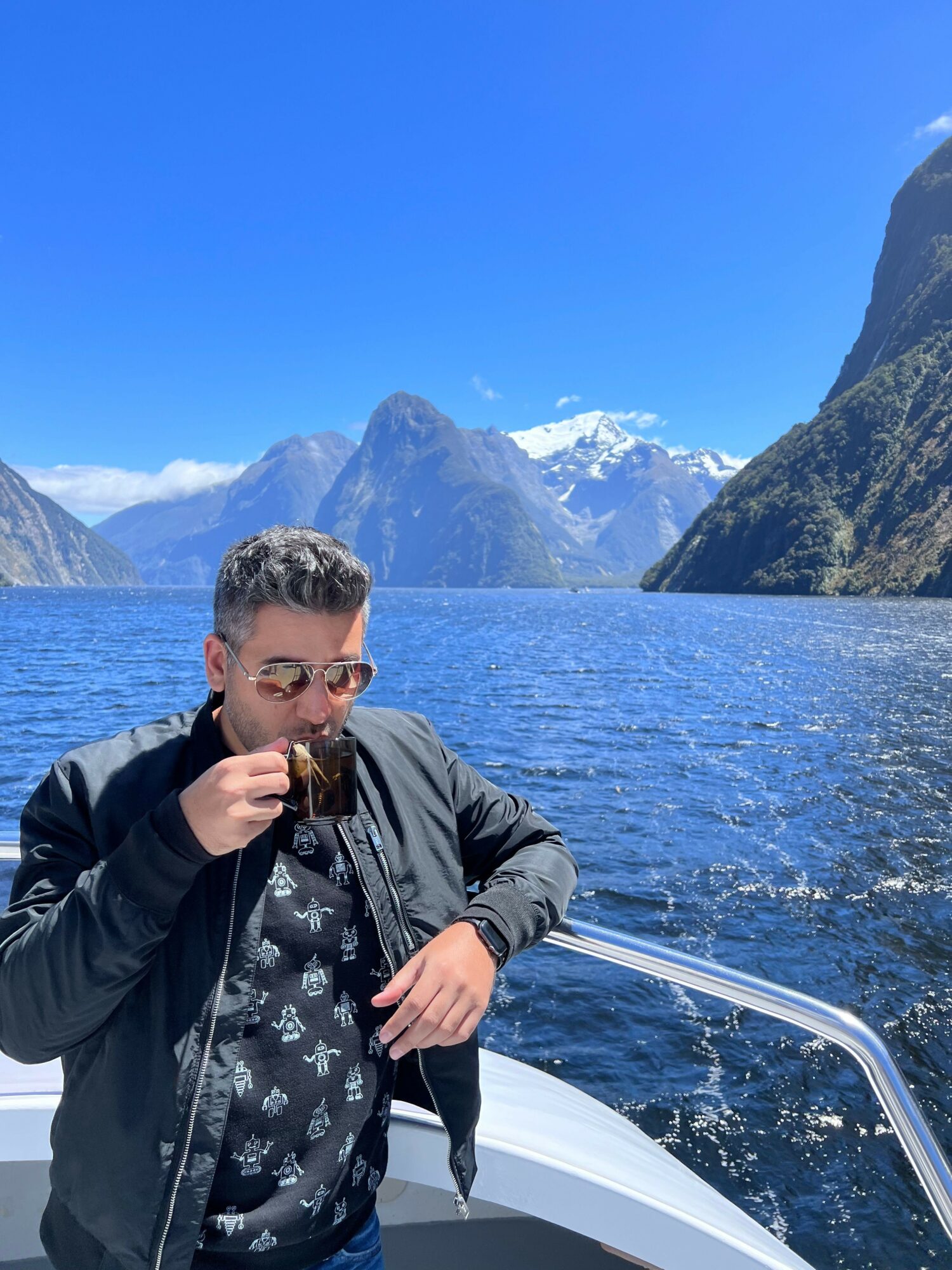 Man wearing sunglasses and a jacket, drinking from a cup on a boat with a mountain lake background.