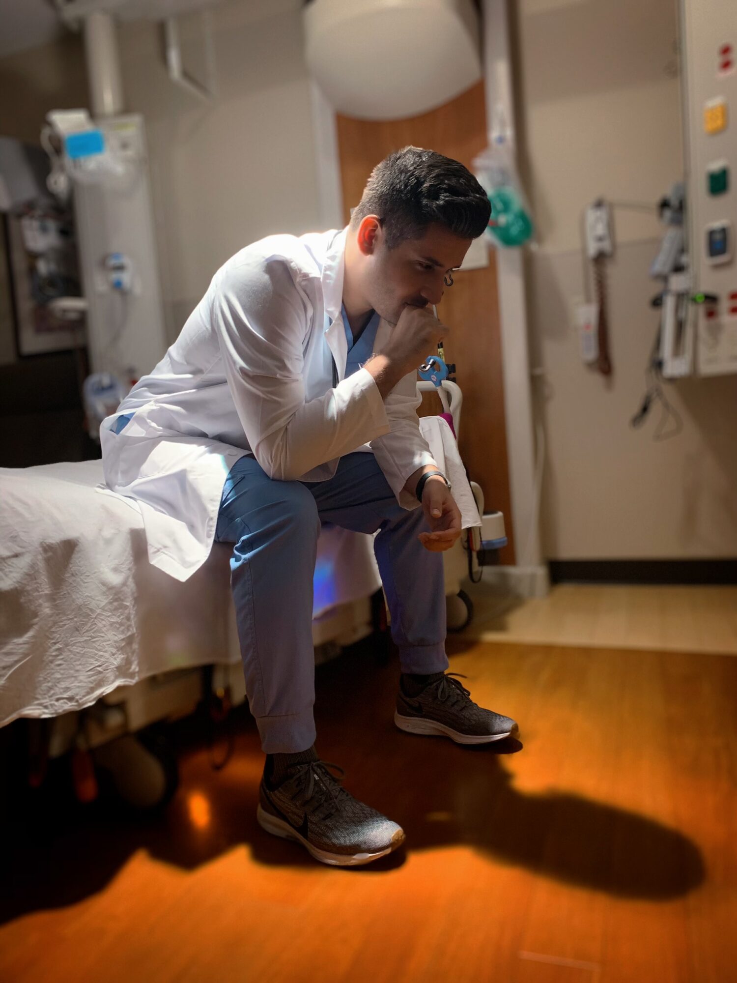 Young man sitting on hospital bed, leaning forward with hand near mouth, in a medical room with equipment on walls.