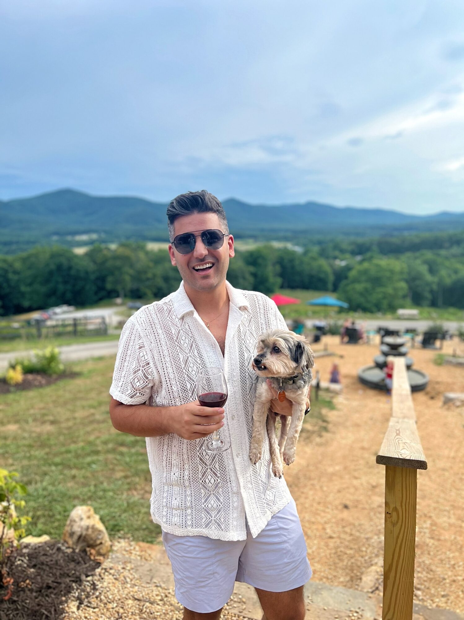 Man holding a small dog and a glass of wine outdoors with mountains and sky in background