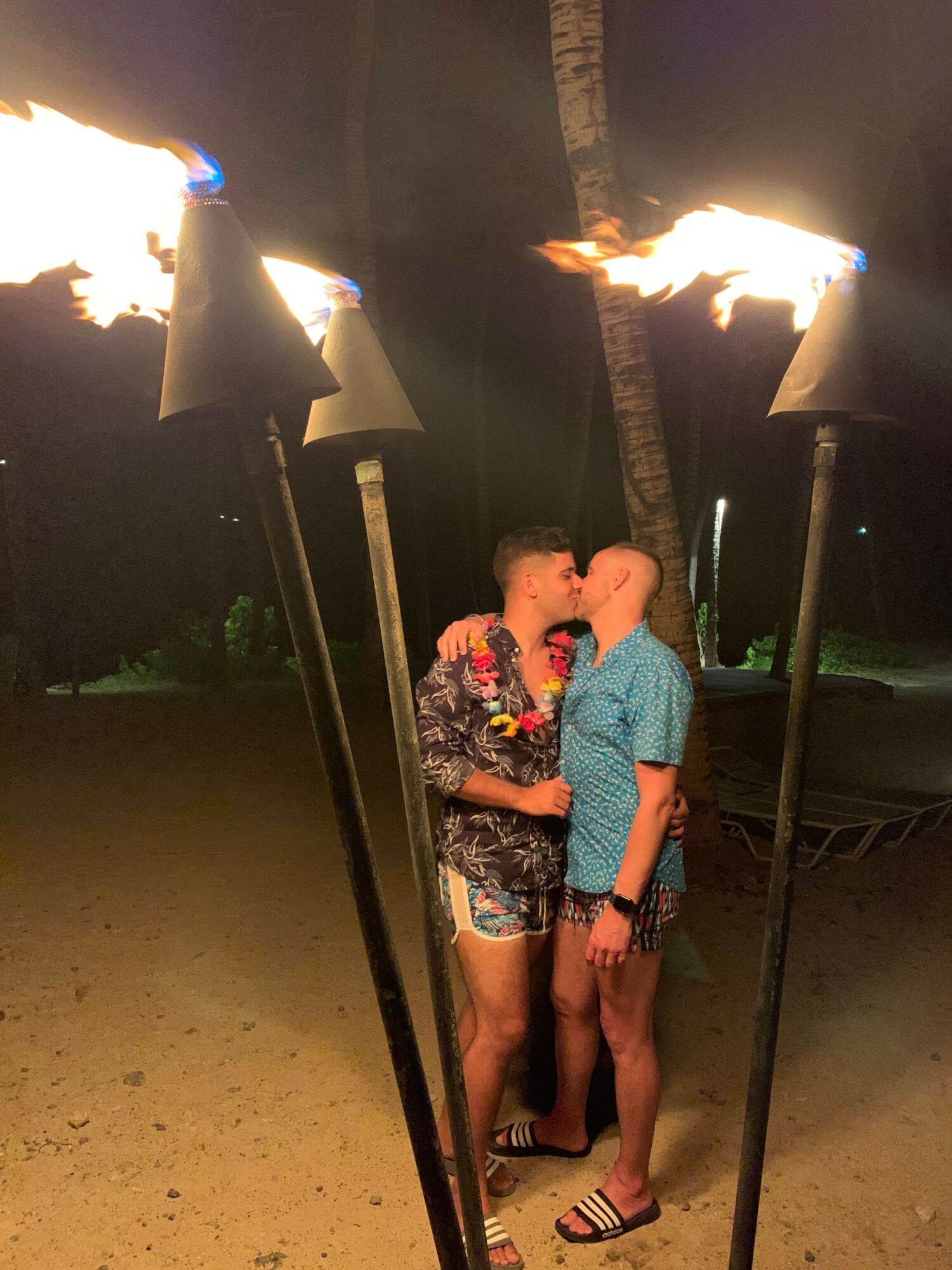 Two people kiss outdoors at night surrounded by tiki torches with flames, trees in background, one wearing a floral shirt, the other in a blue shirt.