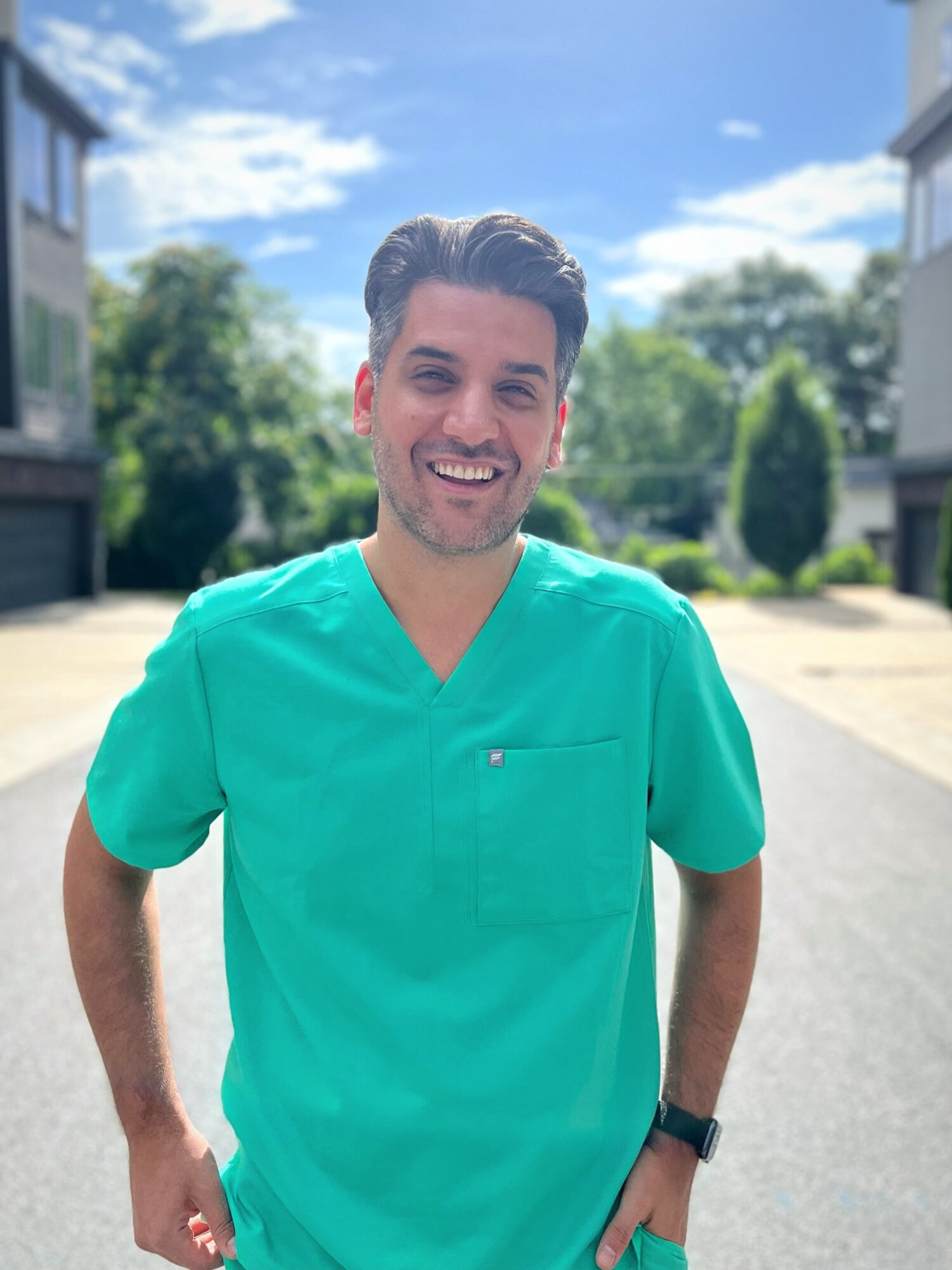 Young man in teal medical scrubs smiling outdoors on a sunny day.