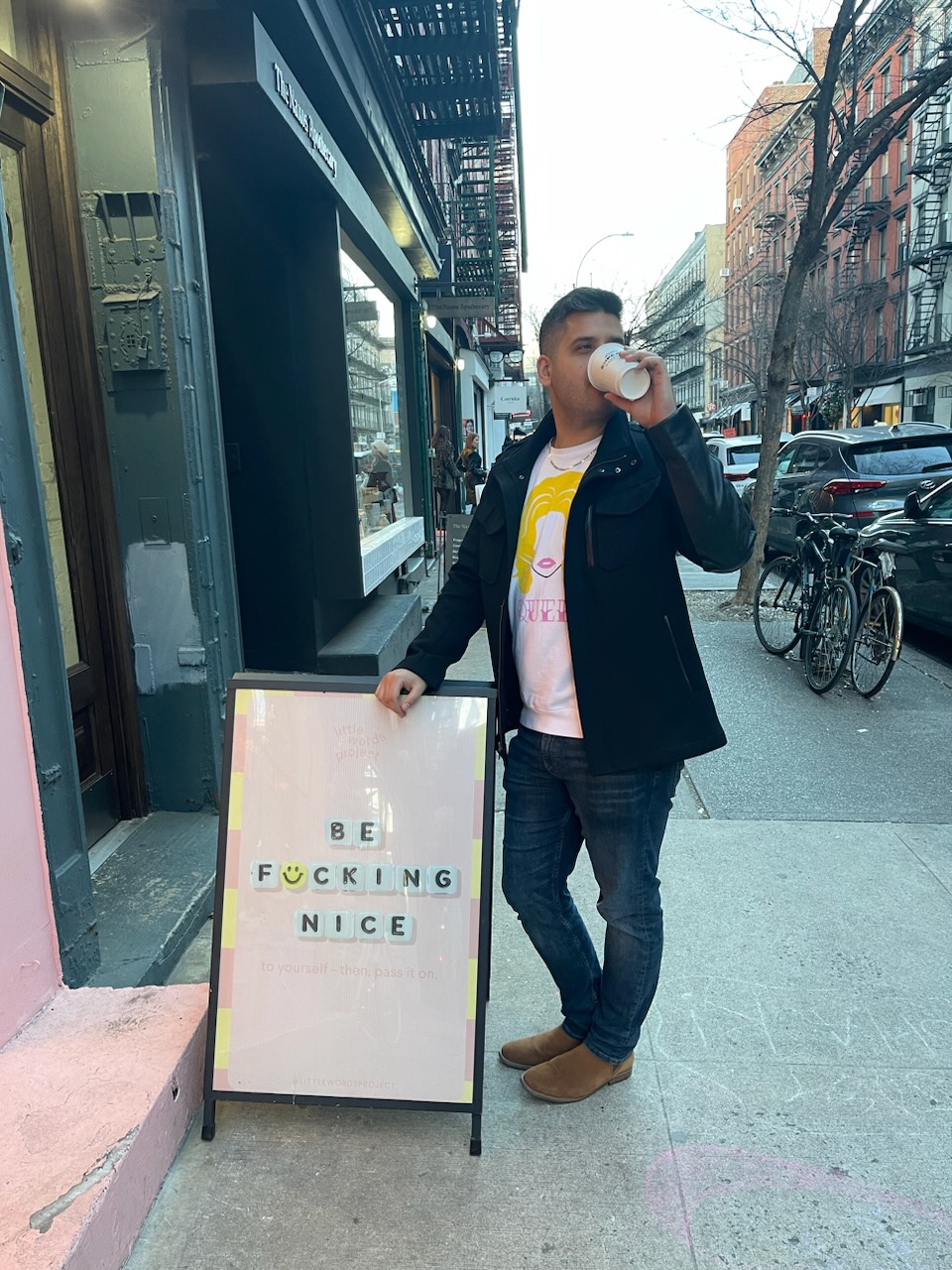 Person standing on sidewalk drinking from cup, next to sign with message, city street with bicycles and buildings in background.