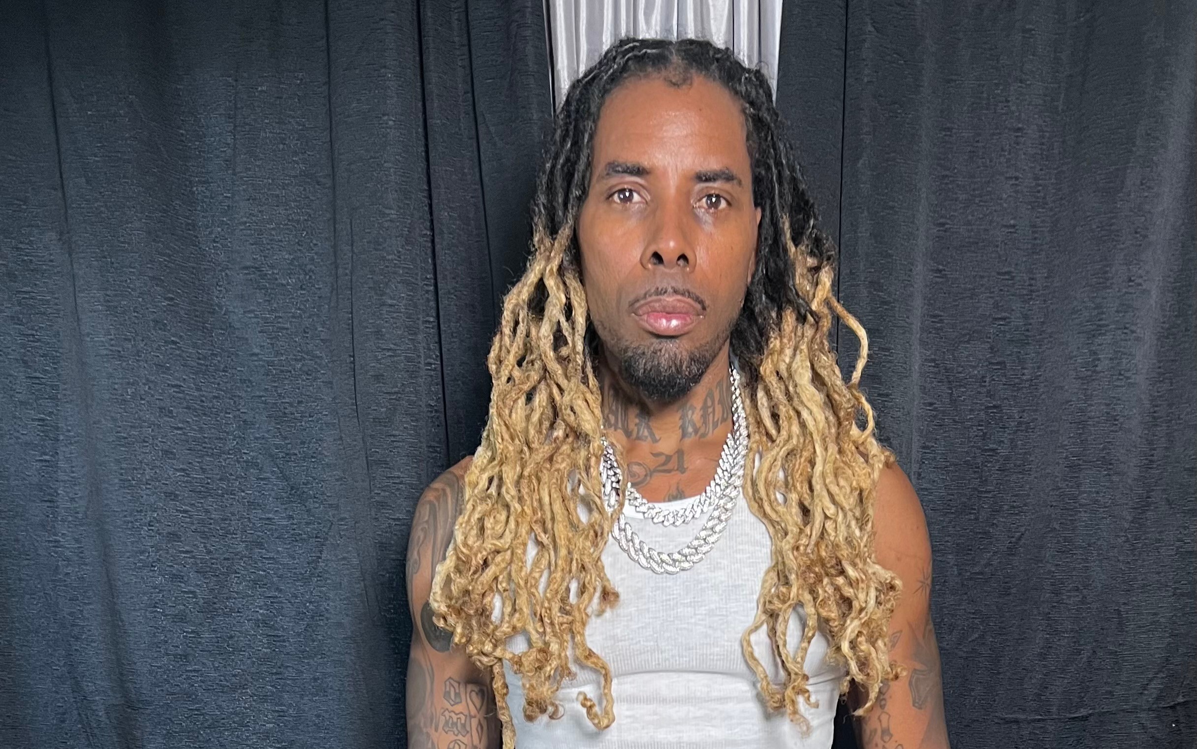 Man with long, blonde dreadlocks wearing a white sleeveless shirt stands in front of dark curtains.