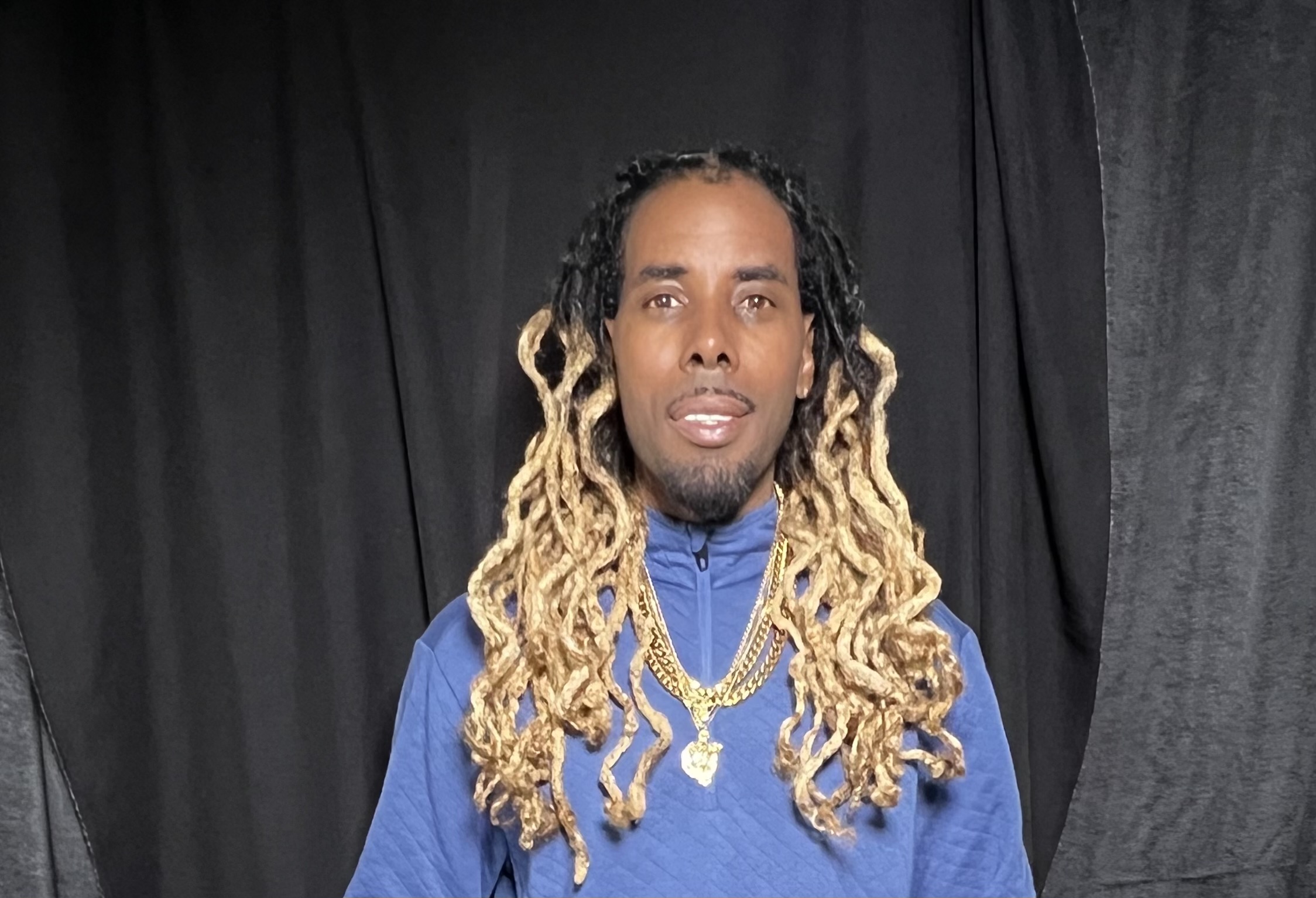 Man with long, blonde dreadlocks wearing a blue jacket and gold chains, standing in front of black curtains.