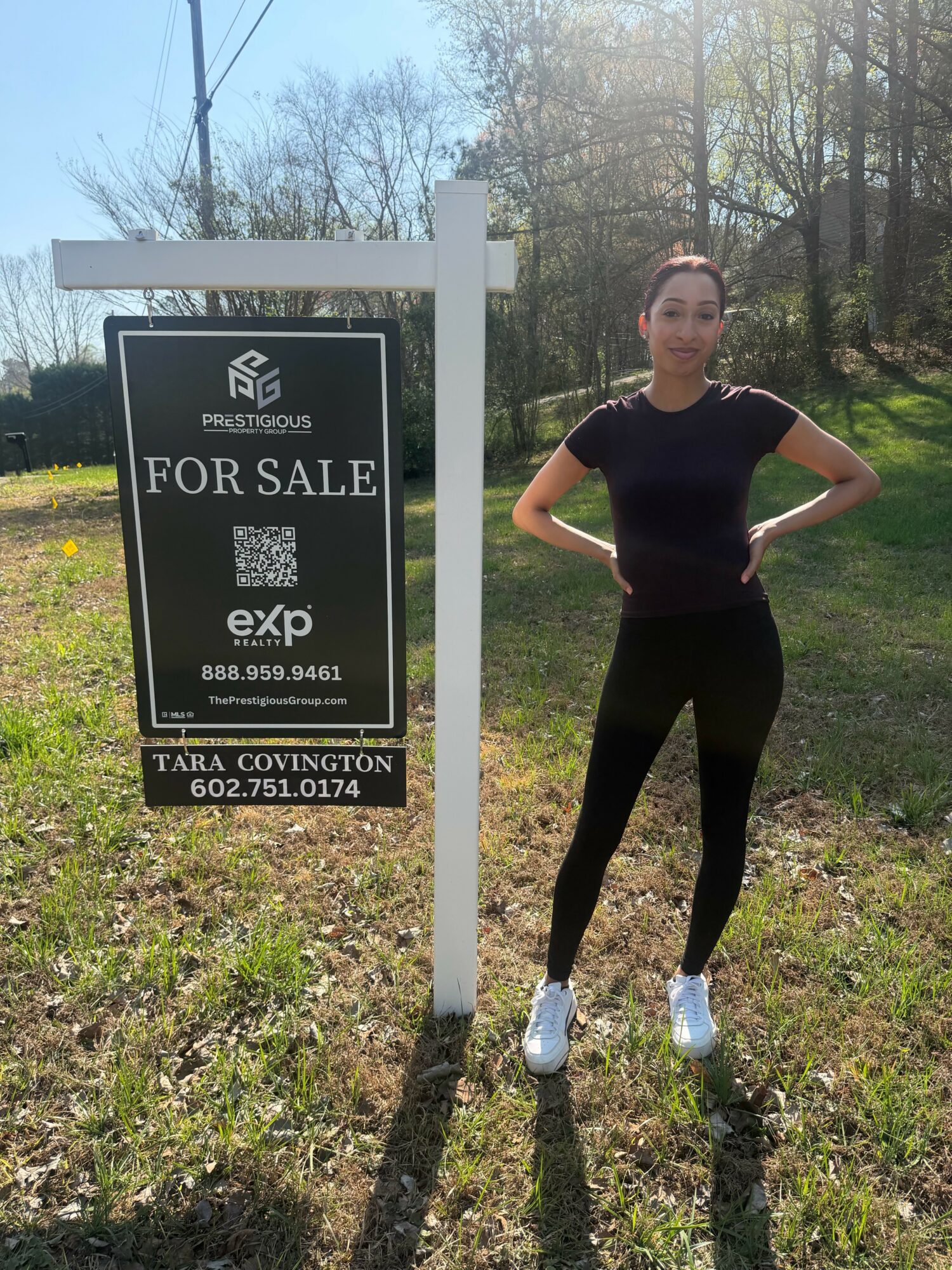 Woman standing outdoors next to a for sale sign, with trees and sunlight in the background.