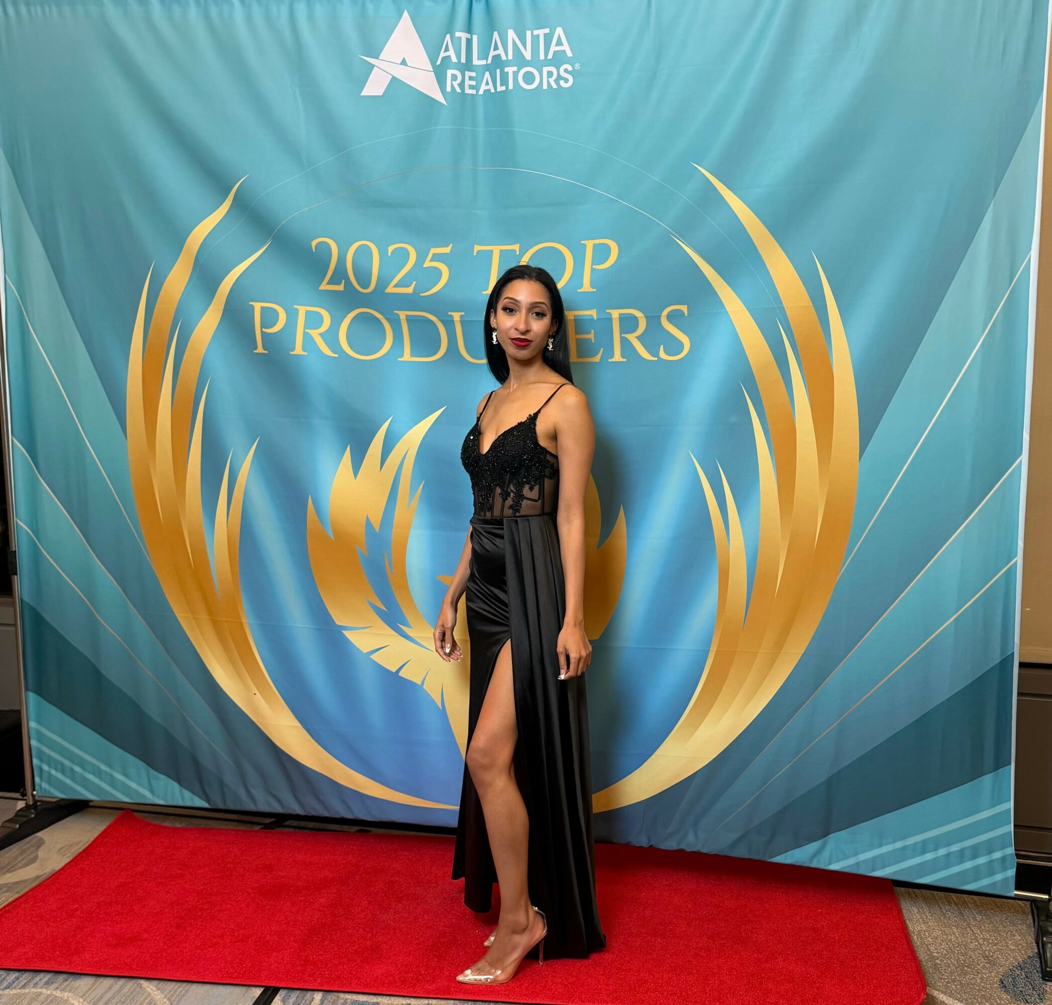 Woman in black gown standing on red carpet in front of a blue backdrop with gold design and text.
