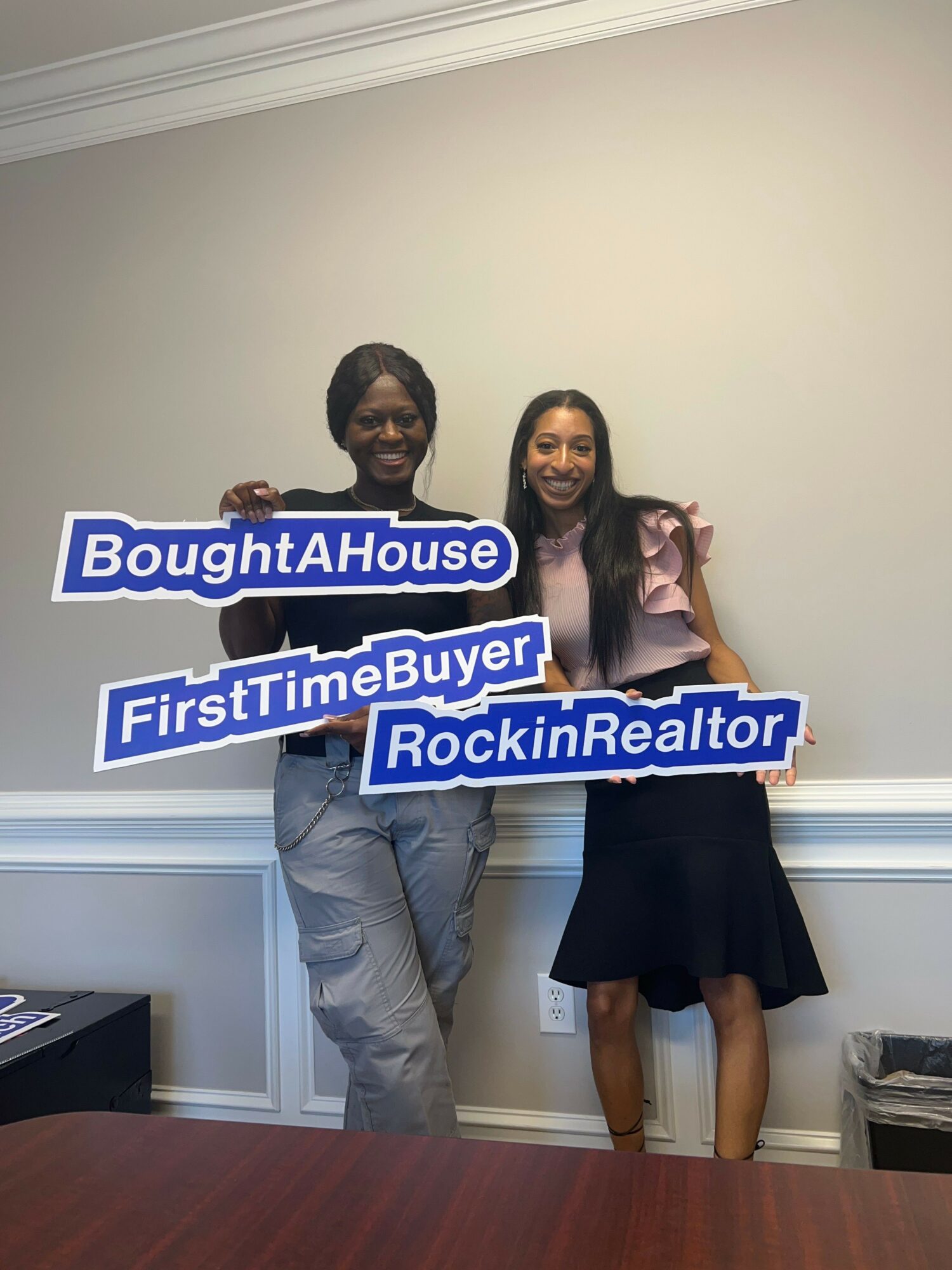 Two women standing indoors, holding signs that say 'BoughtAHouse,' 'FirstTimeBuyer,' and 'RockinRealtor,' smiling at the camera.