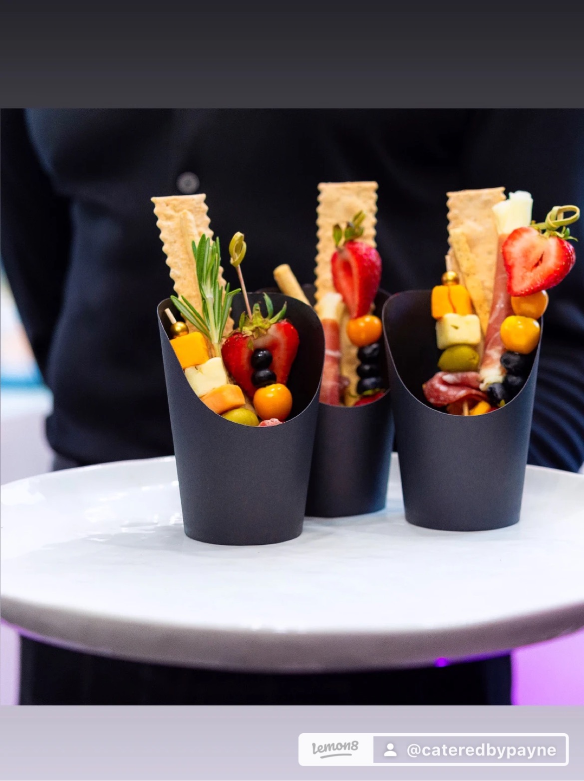 Three black cups filled with fruit and crackers on a white surface, with a person in dark clothing in background.