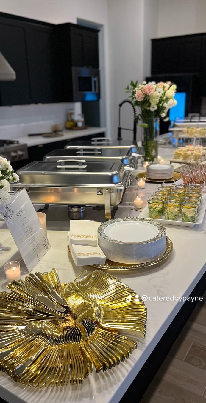 Buffet table with chafing dishes, floral arrangements, and gold decorative items in a modern kitchen setting.