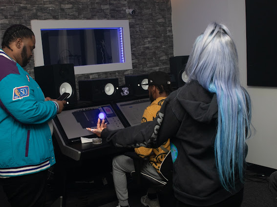 Three people in a music studio with equipment, one with long blue hair, using a touchscreen device.